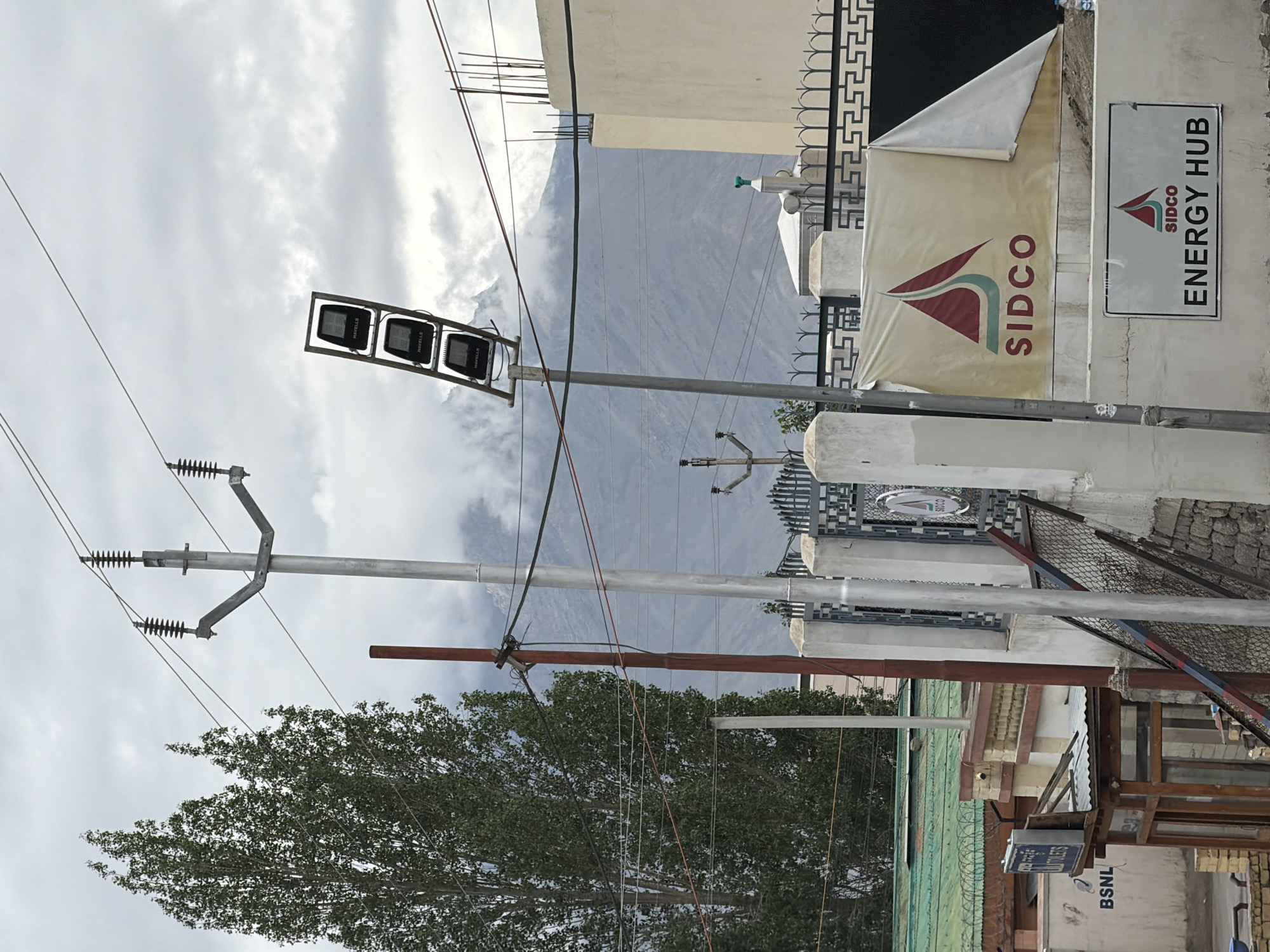 The image, taken in the city of Leh, India, depicts an outdoor scene dominated by utility poles and wires against a cloudy sky. In the foreground, a tall, slender tree with dense green foliage stands to the left. Several utility poles, made of metal and wood, are visible, supporting a network of power lines that crisscross the frame. One pole has a cluster of three prominent, dark-colored floodlights mounted on it.
A white wall with a decorative black and white patterned frieze runs across the middle of the image. On this wall, a large banner with the SIDCO logo is partially unfurled, revealing the text "SIDCO" below a red and teal graphic. Below the banner, a smaller sign also displays the SIDCO logo and the text "ENERGY HUB". To the left of these signs, a building with a green corrugated roof is partially visible, along with a smaller structure featuring a wooden-framed opening and a blue sign that reads "TOILETS". A "BSNL" logo is also present on this structure.
Further left, a reddish-brown pole stands near the base of the large tree, also supporting wires. In the background, a mountain range, partially obscured by clouds, forms the backdrop. The overall environment appears to be an urban or semi-urban area with infrastructure for power distribution and visible commercial signage. No people are present in the image.