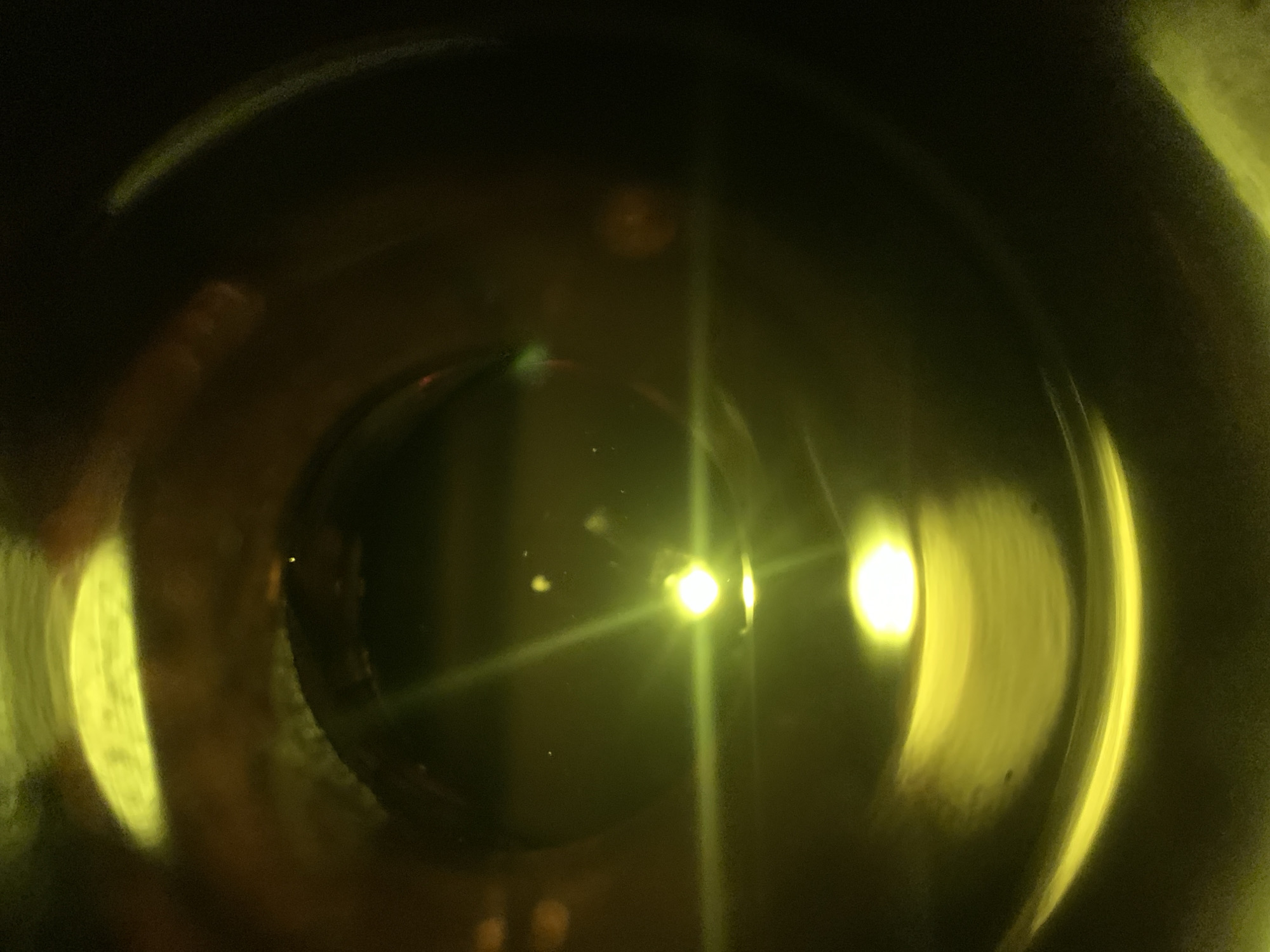 The image appears to be a close-up, somewhat distorted view of liquid within a clear container. Bright light sources reflect off the surface of the liquid and the interior of the container, creating flares and highlights. The liquid itself has a dark color, and the container appears to be round or spherical. The setting within the city of New Stadium, Nigeria, is not visible in the image.