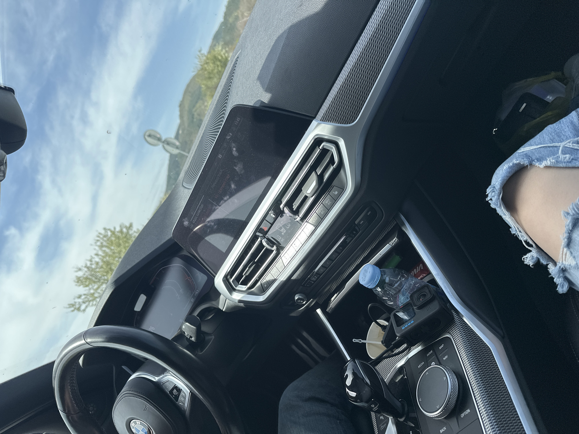 The image provides an interior view of a modern car, specifically a BMW due to the visible steering wheel logo and dashboard elements, taken from the passenger's perspective. The legs of a person wearing distressed denim shorts are prominent in the foreground on the passenger side.
Through the windshield, a bright blue sky with scattered white clouds is visible, along with distant green hills or mountains, indicating clear and sunny daytime weather. A street light or sign pole can also be seen outside. The digital instrument cluster displays "D7 +16.5" and "76."
The central dashboard features a large infotainment screen showing "11:45" as the time, along with "Music collection," "Previous track," and "Next track." Below it are silver-accented air vents and climate control buttons, including "MAX" and "A/C." The center console between the seats holds a "VIVA" brand water bottle, a "GoPro" camera, a white disposable cup, and a charging cable. The gear selector area displays "P," "R," and "OFF," while the iDrive controller is surrounded by buttons labeled "MEDIA," "HOME," "MAP," "NAV," "OPTION," and "BACK." The car's interior trim includes a textured, possibly carbon-fiber-like pattern on the dashboard and console. The scene suggests a casual journey or stop in Madrets, Bulgaria, with the vehicle either parked or slowly moving.