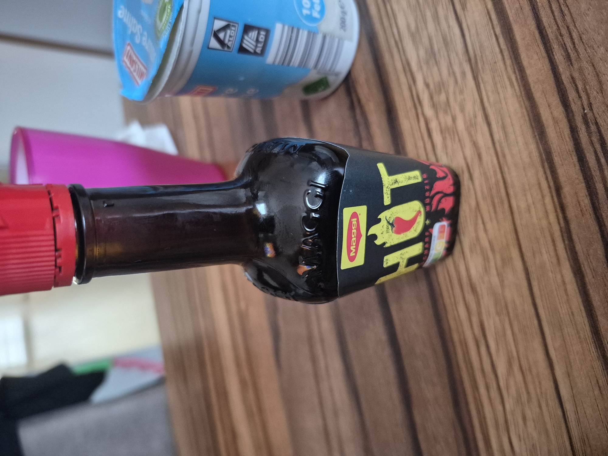 The image shows a close-up of a bottle of Maggi Hot sauce and a container of Aldi dairy product on a wooden table. The bottle of Maggi Hot sauce is dark brown glass with a red cap and a black label featuring the word "HOT" in large yellow lettering with a red chili pepper design. The Aldi container is white with a blue lid and features the Aldi logo. In the background, a pink plastic cup is visible, and a blurry figure wearing black and green clothing is present. The scene is indoors, and the lighting suggests it is daytime. There is no specific indication of the location in Barbing, Germany.