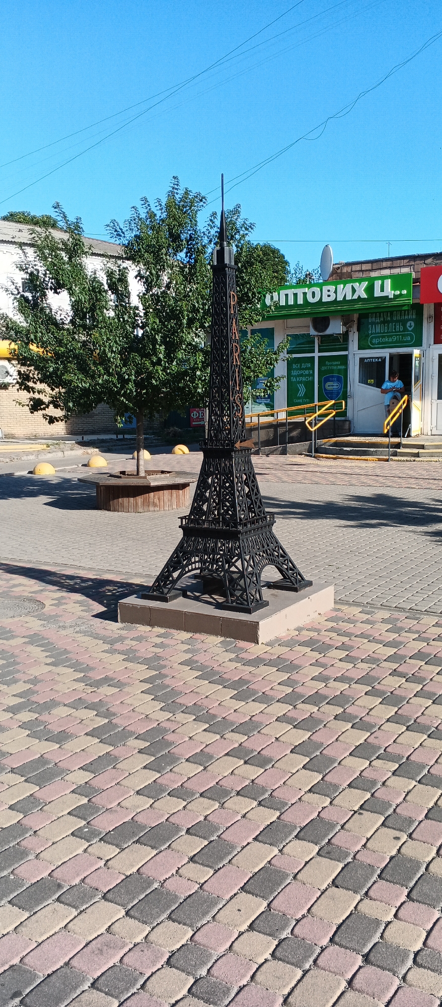 The image shows a miniature Eiffel Tower replica placed on a paved area in front of a commercial building. The Eiffel Tower is black and made of metal lattice. It is mounted on a light-colored concrete base. To the left of the replica is a green tree with a wooden bench around its base. In the background, there is a building with a green awning that reads "ОПТОВИХ Ц..". The building appears to be a store or pharmacy, with a sign that says "АПТЕКА". A person wearing a blue shirt is standing in the doorway of the building. The ground is paved with colorful interlocking bricks. Yellow bollards are visible in the foreground. The sky is clear and blue. This scene is located in the city of Smila, Ukraine, on a sunny day.