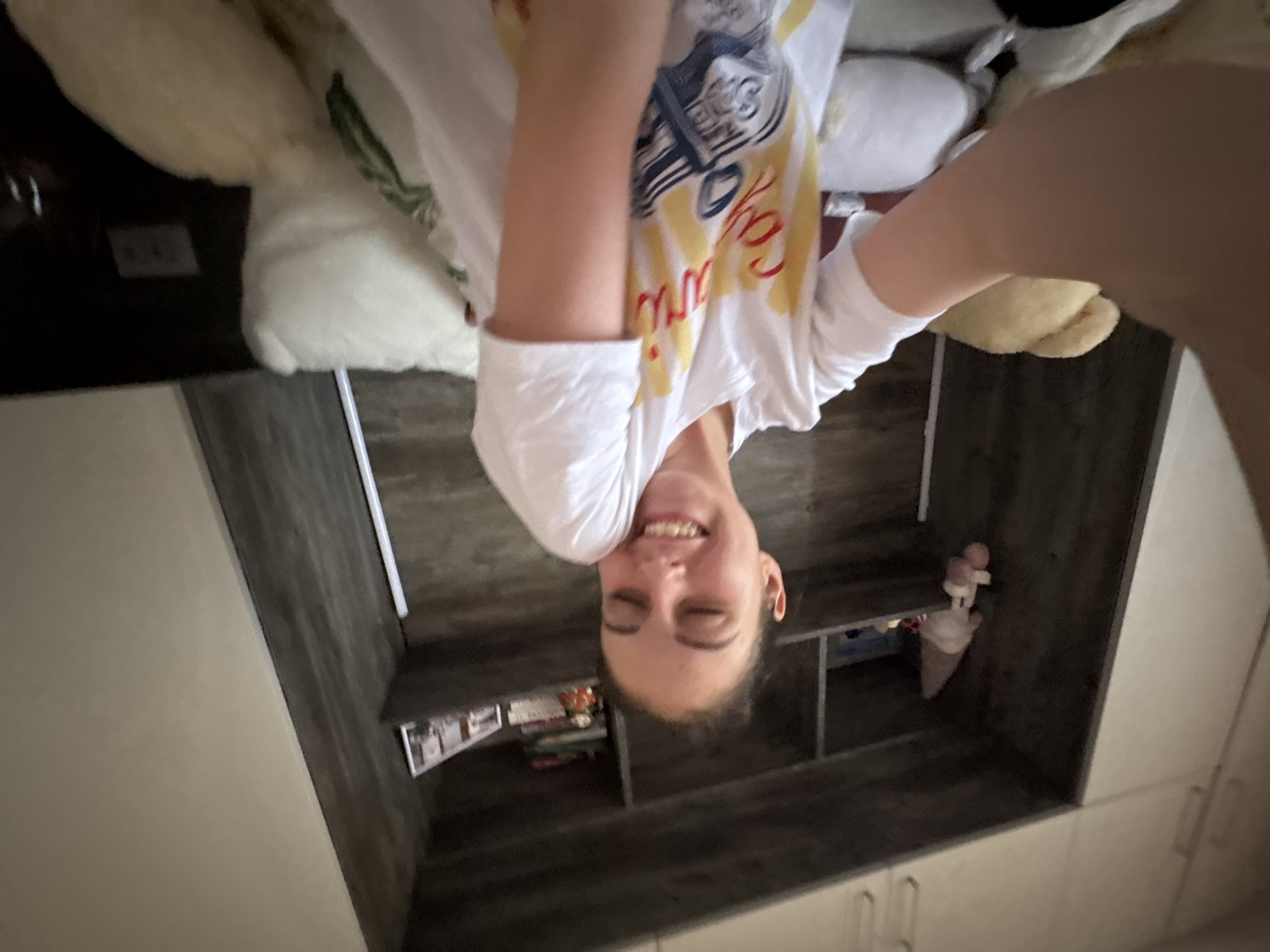 A person is upside down in an indoor setting. The person is wearing a white t-shirt with a graphic print. Behind the person is a set of shelves containing various items, including books and a toy ice cream cone. The background appears to be a room with light-colored walls and a dark gray shelf unit. The location is not discernible from the image.