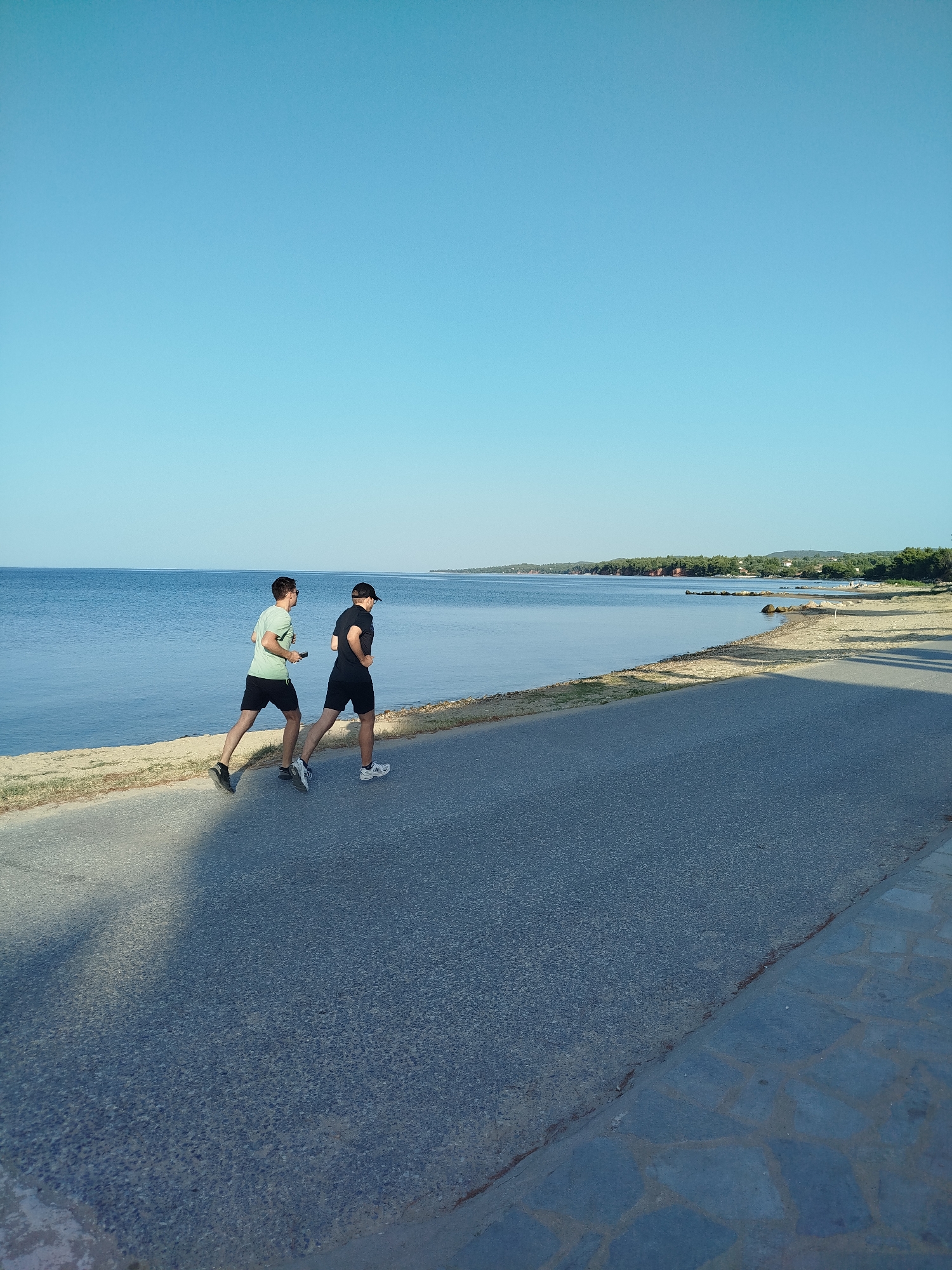 Two young men are jogging side-by-side on a paved path along a shoreline in Sithonia Municipal Unit, Greece. The man on the left wears a light green t-shirt and black shorts, while the man on the right wears a black t-shirt, black shorts, and a black baseball cap. They are running from left to right, away from the camera. The path is bordered by a sandy beach and then the calm blue sea. In the distance, the coastline is lined with trees and some buildings. The sky is clear and blue. The lighting suggests it is late afternoon or early morning.