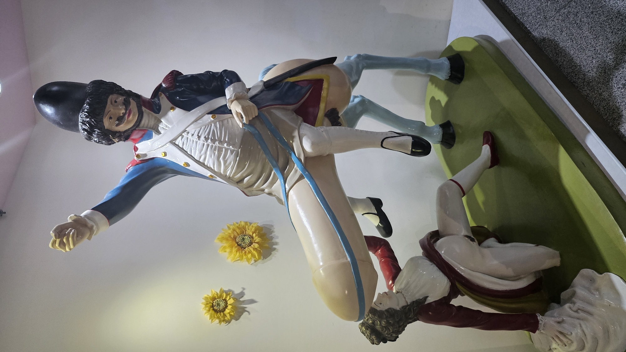 This image depicts a surreal sculpture on display in Seogwipo-si, South Korea. The sculpture features a man dressed in a historical military uniform, complete with a tall, black hat and a prominent mustache, riding a horse. However, the horse's anatomy is distorted, with a phallic shape where its hindquarters and body should be. Beneath the horse, a figure in a red coat and white attire is positioned, appearing to be interacting with the phallic element of the horse. Two artificial sunflowers are mounted on the white wall behind the sculpture. The sculpture is set on a green base, which is itself placed on a light-colored display platform. The background consists of a plain white wall.