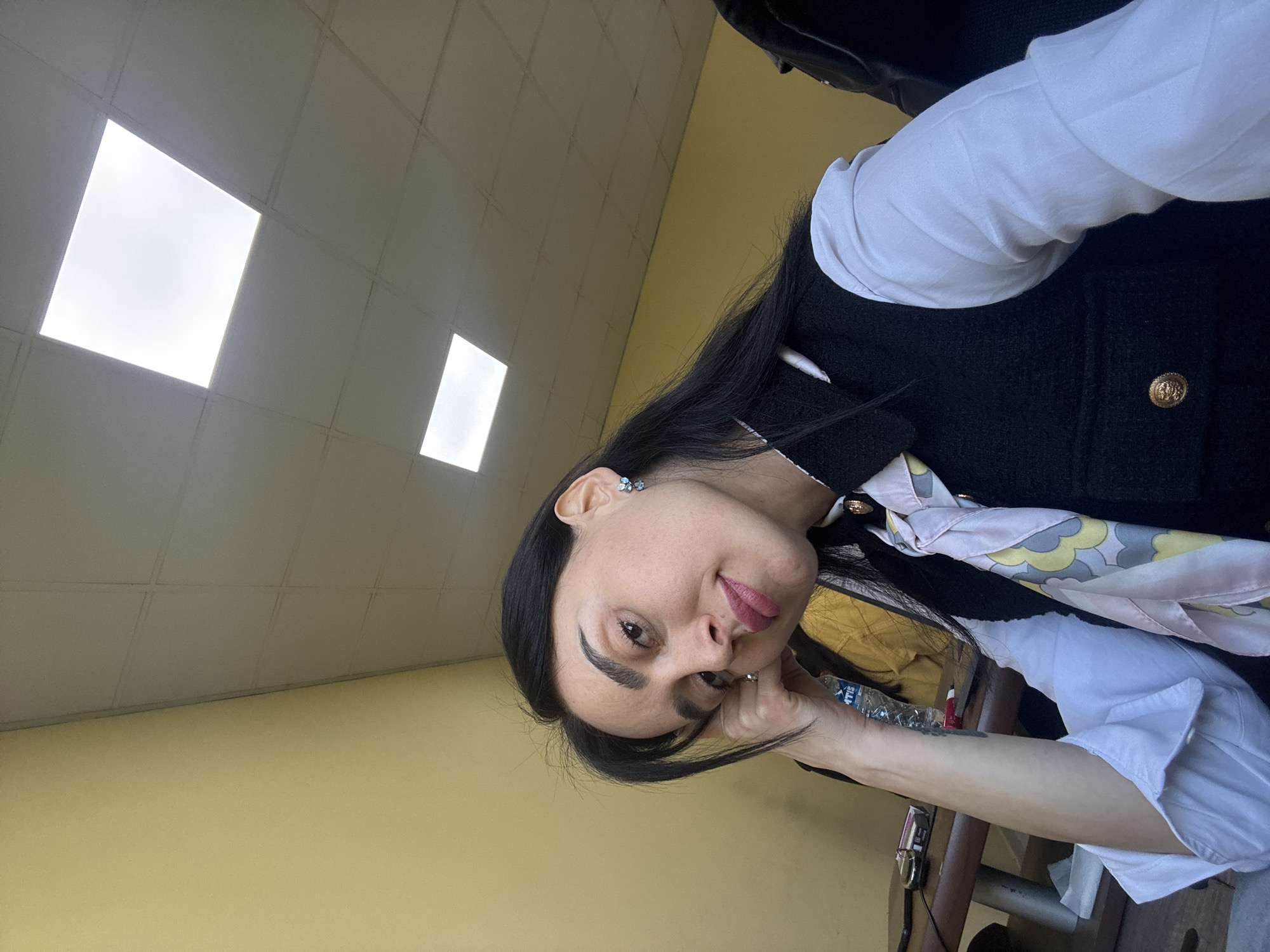 An indoor portrait displays a woman in what appears to be an office or public building in Tbilisi, Georgia. The image is oriented sideways, with the woman's head tilted towards her left shoulder, looking calmly at the camera with a subtle smile. She has long, straight dark hair and distinct dark eyebrows, with subtle eye makeup and pink lipstick. A small, light blue butterfly or flower-shaped earring adorns her visible left ear.
She is dressed in a white long-sleeved shirt layered under a dark, possibly black, textured jacket or vest, featuring a prominent gold decorative button on the lapel. A light-colored scarf with abstract, swirling patterns in shades of pink, yellow, grey, and white is tied around her neck. Her left hand rests gently near her chin, and she wears a ring on her right ring finger.
The background shows a plain light yellow-beige wall, meeting a white tiled ceiling that contains several square, brightly lit fluorescent light panels. These panels are the primary source of illumination. Partially visible on a surface next to her arm are a plastic water bottle with some faint, unreadable text, and the red tip of what appears to be a pen or marker, along with other indistinct items. The overall setting suggests a typical indoor environment.