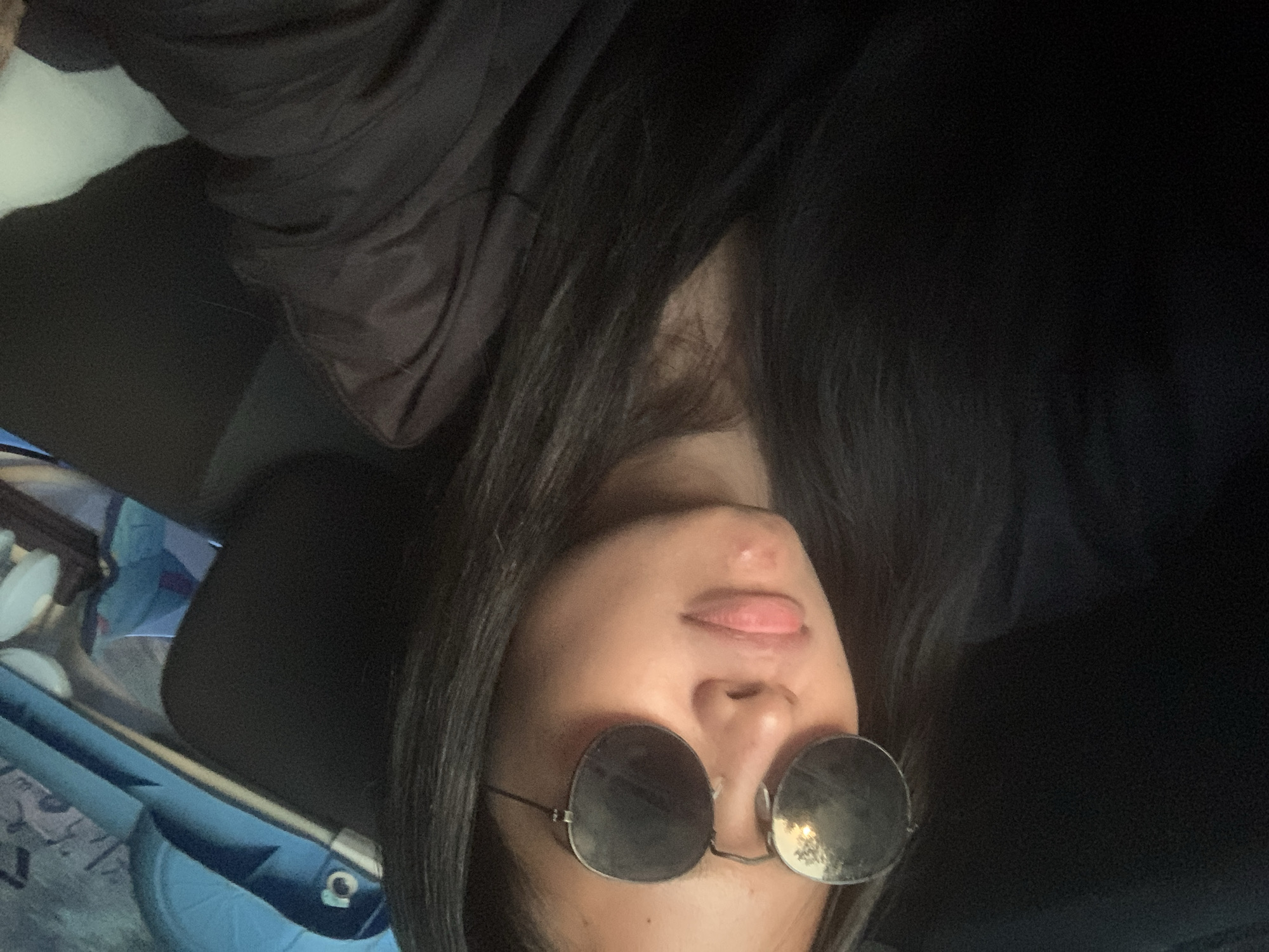 A person with long dark hair is wearing round sunglasses and a black outfit. The individual's face is tilted upwards, and their tongue is slightly protruding. To the left, there are glimpses of a blue bag and other unidentified objects. It is not possible to determine if the location is in Batu Batu, Indonesia, from the provided image.