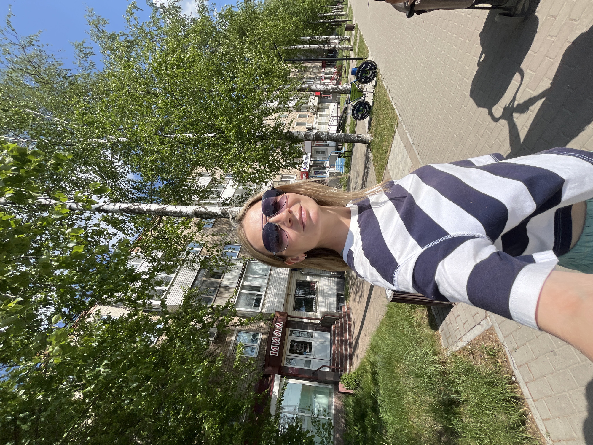 A woman with blonde hair and sunglasses stands on a brick-paved walkway in Chaikovsky, Russia, holding a camera and taking a selfie. She wears a blue and white striped long-sleeved shirt. Behind her is a row of birch trees with green leaves. Buildings are visible in the background, including one with the word "МИЛА" (MILA) above the entrance. A street sign with circular hanging panels is also present. The shadow of a stroller is visible on the walkway. The day appears to be sunny.