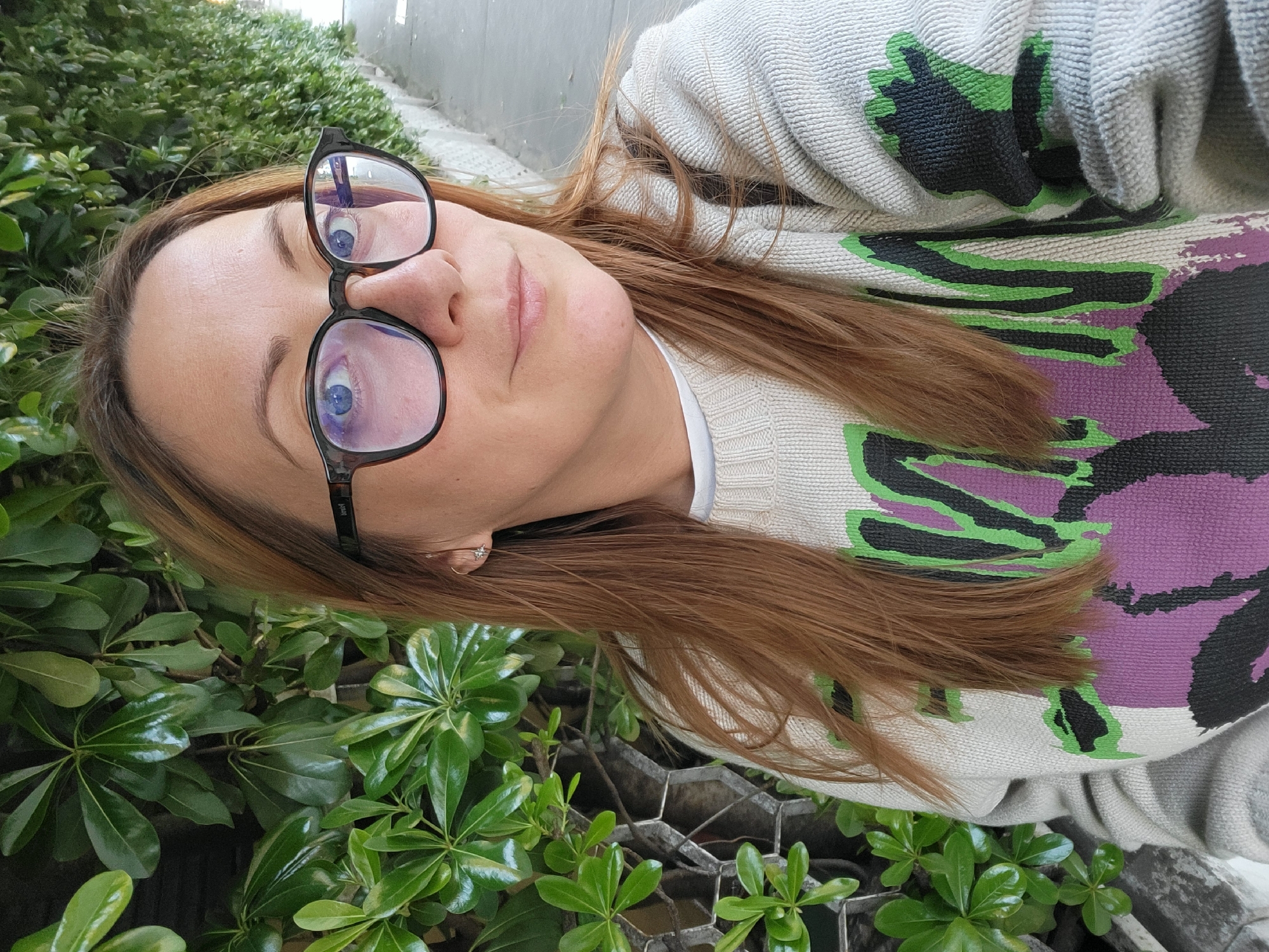 A close-up, portrait-style photograph depicts a woman outdoors, possibly in Budva, Montenegro. The image is composed at an upward angle, showing the woman from the chest up. She has medium-length, reddish-brown hair and striking blue eyes, which are enhanced by the dark, tortoise-shell framed eyeglasses she wears. The brand "POLAROID" is faintly visible on the arm of her glasses. Her gaze is direct and her expression is neutral.
She is dressed in a light grey or off-white knitted sweater, adorned with a distinctive, abstract pattern in green, black, and purple. A white collar or undershirt is visible beneath the sweater. A small, star-shaped stud earring can be seen in her right ear.
The background is filled with lush, vibrant green foliage, characteristic of dense bushes or hedges, suggesting a garden or landscaped area. In the upper background, a light-colored wall or building is discernible, while grey, possibly metal, structures (like planters or fencing) are partially visible in the lower background. The bright and clear lighting indicates that the photo was taken during the daytime.
