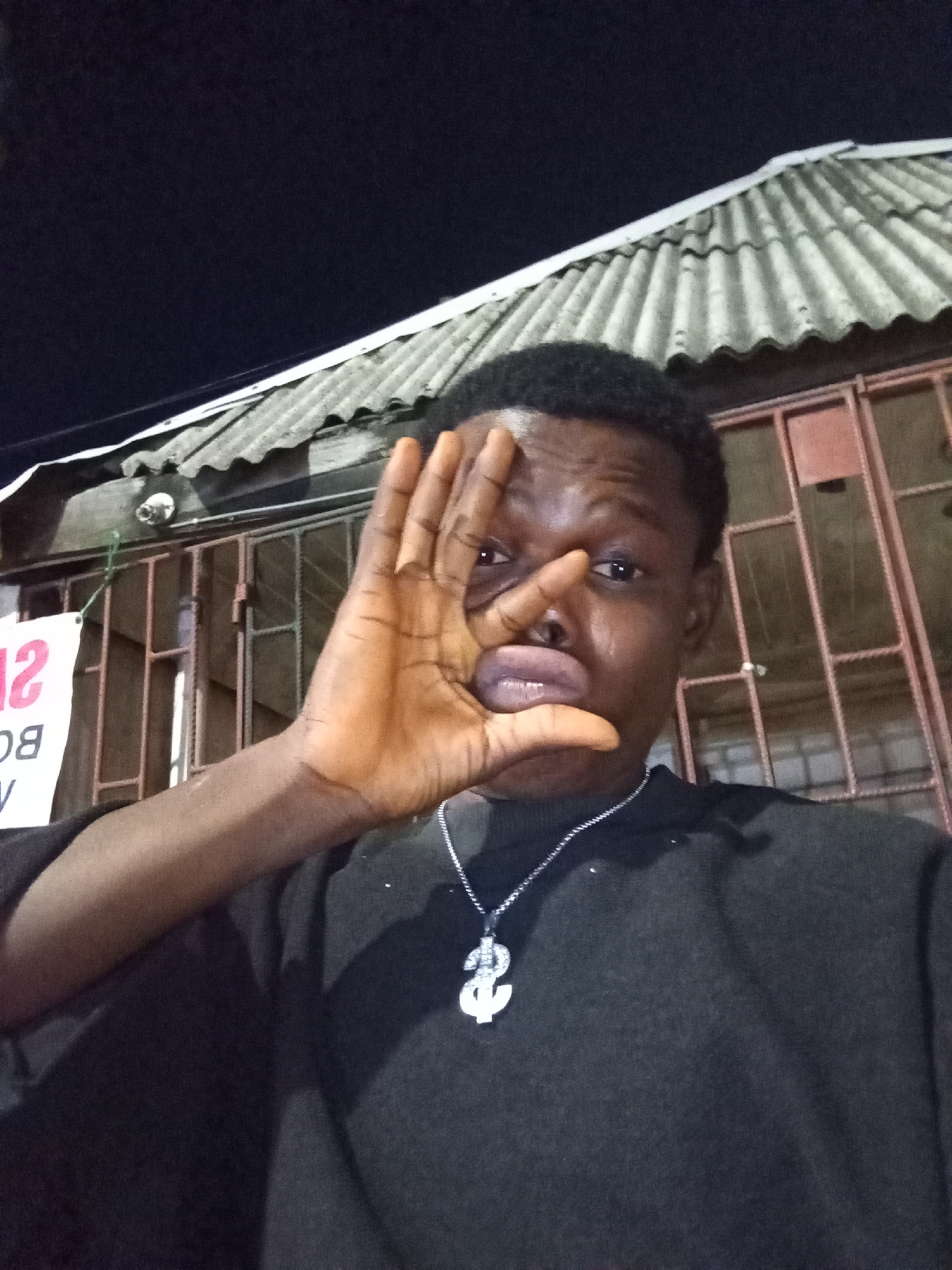 A selfie shows a dark-skinned young man with short black hair, wearing a black sweater and a silver chain with a dollar-sign pendant. He is holding his right hand up to his face, partially obscuring his left eye and nose. The background consists of a building with corrugated metal roofing. Behind the man are metal bars possibly indicating a window or door. A partial sign with the number "12" is visible on the left. The setting appears to be outdoors at night in the city of Ado, Nigeria.