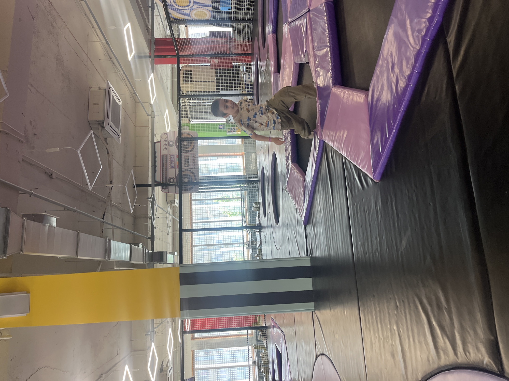 A young boy is playing on a trampoline course inside a facility, identifiable by its interior structure featuring overhead ductwork, fluorescent lighting fixtures, and large windows that provide natural light and allow views of the cityscape outside, including buildings and green foliage. The trampoline course has black surfaces with sections of padded purple structures that the boy is maneuvering around. The boy wears a printed t-shirt and khaki pants and appears to be in motion, suggesting active play. A large wall graphic of a boombox is visible in the background. Based on provided information, it is located in the city of Kaspiysk, Russia.