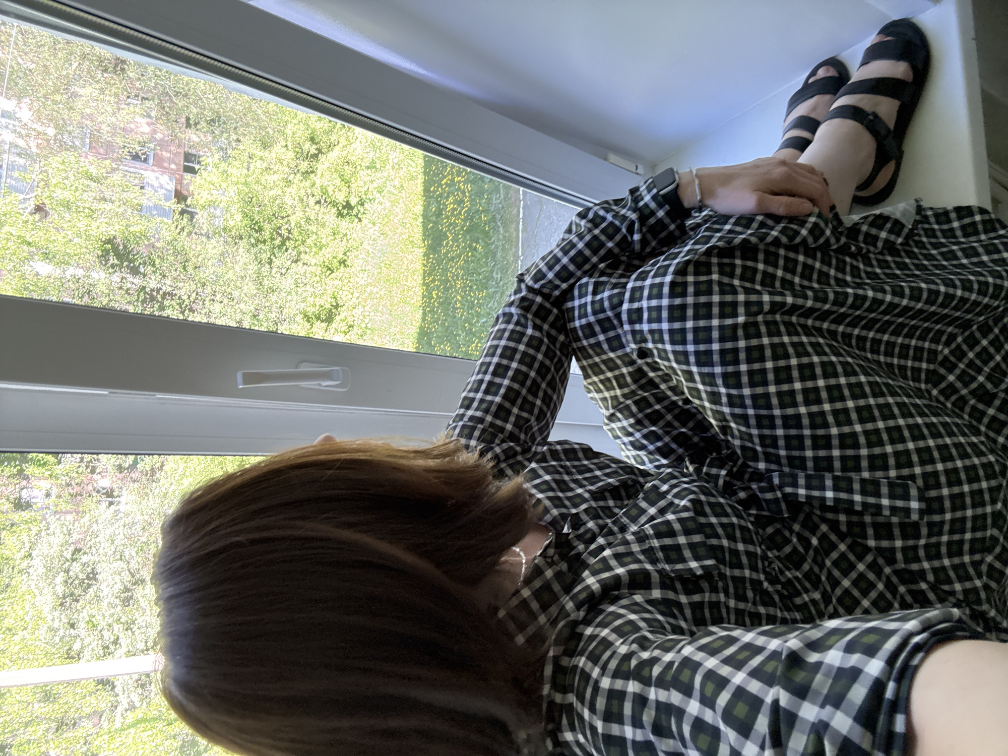 A woman lies in a window seat looking out onto a tree-filled landscape. She is wearing a green and white checkered long-sleeved garment. Her brown hair is visible from the back. She is wearing black sandals. Her wrist is adorned with an Apple Watch and a silver bracelet. The view outside the window features lush green trees and what appears to be a residential building in the distance. Based on the metadata, the image was taken in Chaikovsky, Russia.