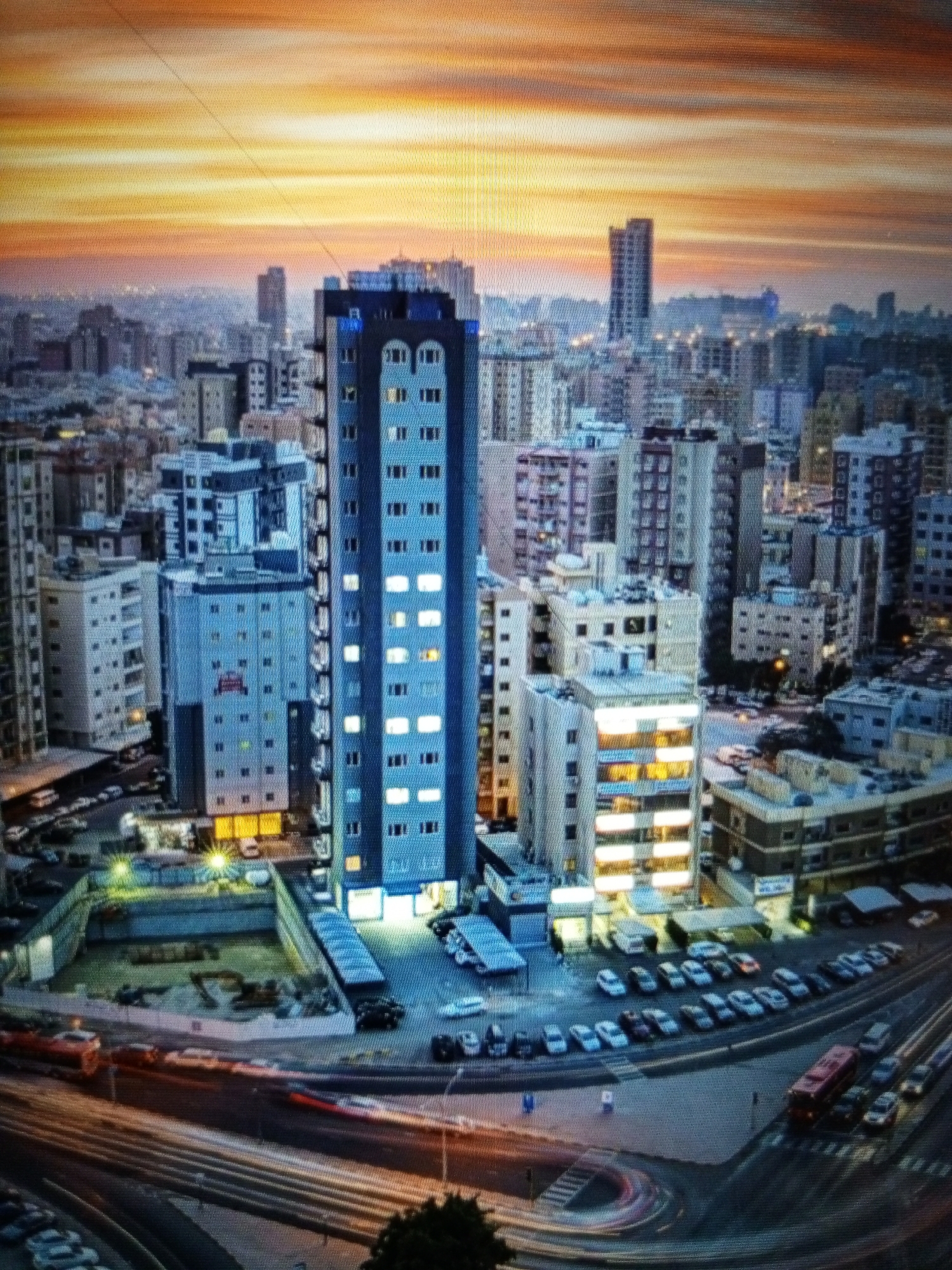 This is a high-angle, full-shot photograph of a city skyline at dusk. The sky is filled with streaks of orange, yellow, and pink, suggesting a sunset. In the foreground, a tall, slender blue building with many illuminated windows dominates the scene. Surrounding this central building are numerous other urban structures of varying heights and architectural styles, also displaying illuminated windows, indicating the city is populated and active. Several cars are visible on the streets and in parking lots, with some displaying light trails, suggesting movement and a long exposure time for the photograph. The ground level appears to be a mix of roads, sidewalks, and parking areas. The overall impression is of a dense, modern urban environment under a dramatic sky. The image is said to depict the city of Golbembal, Nigeria, although this is not visually verifiable from the image alone.