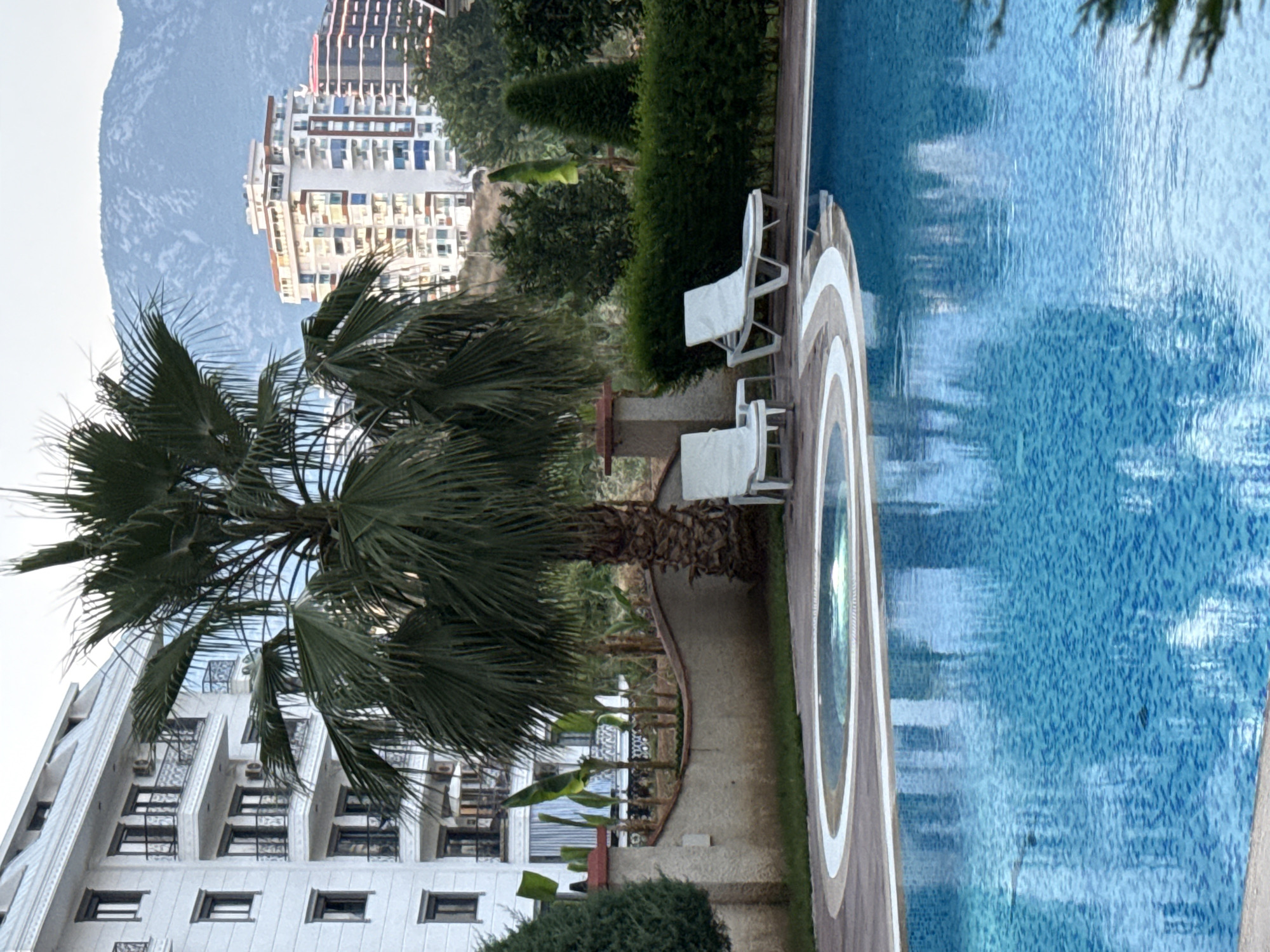 The image depicts an outdoor scene with a swimming pool, a building, and a palm tree. The swimming pool is a prominent feature, filled with blue water and tiled with small blue squares. A wooden deck surrounds a portion of the pool, and a section of the pool has a circular jacuzzi or whirlpool. Two white plastic chairs are placed on the deck near the pool.
To the left of the pool, a modern white building with multiple floors and balconies is visible. The building has many windows and air conditioning units. A large palm tree with its fronds spread out is positioned in front of the building and partially obscures it. In the background, a mountainous landscape and more buildings are discernible.
The image appears to be taken during the day, with soft, diffused light suggesting it might be early morning or late afternoon, or an overcast day. The presence of the palm tree and the architectural style of the building, along with the mention of Alanya, Turkey as location context, suggest a warm climate, likely a resort or hotel setting.
There is no visible text in the image. No people are visible, and no specific activity or event is taking place; it seems to be a static scene of a resort environment. The overall mood is peaceful and tranquil, characteristic of a vacation destination.