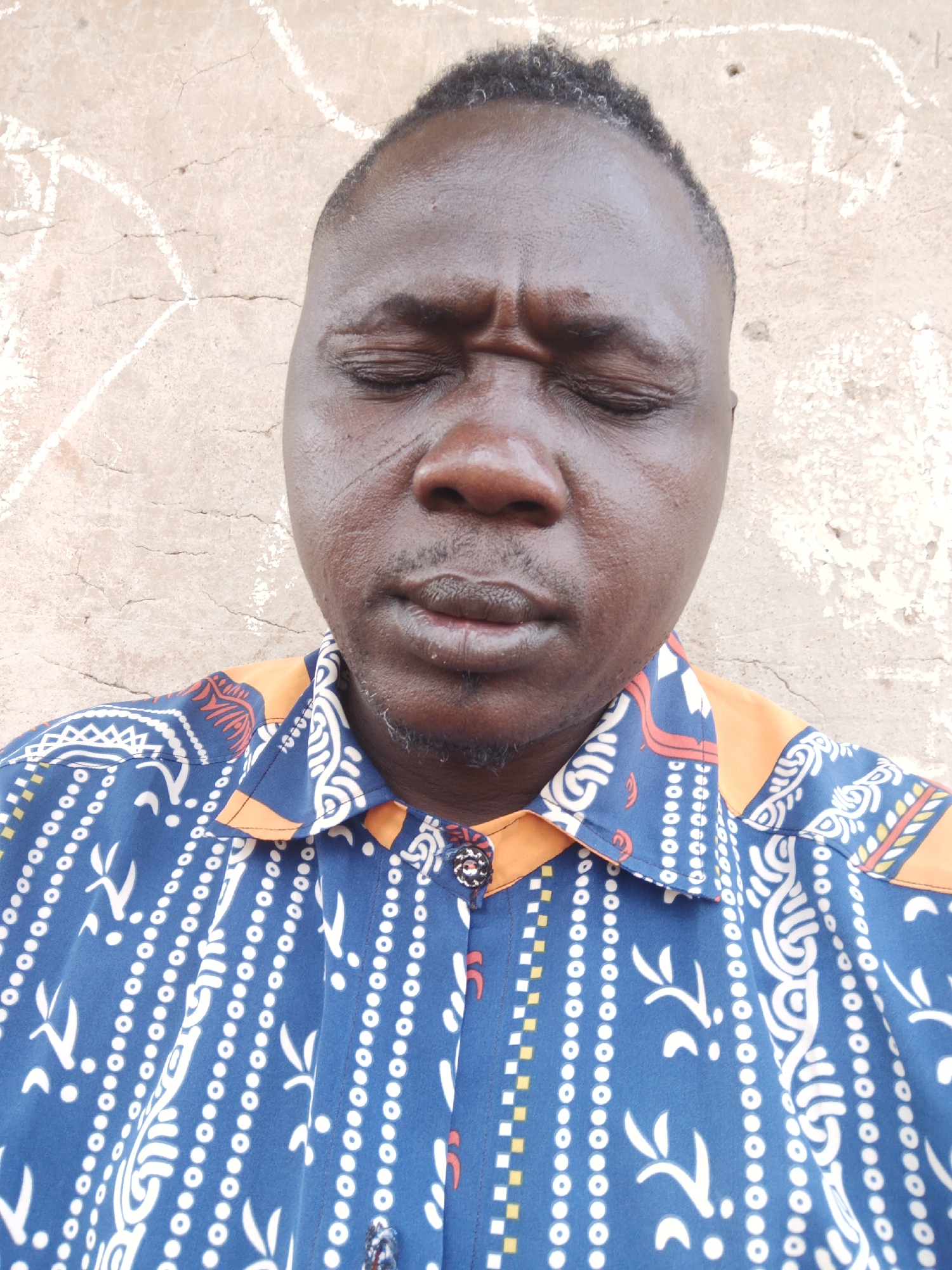 A close-up shot shows a dark-skinned man with closed eyes, furrowed brow, and a somber expression. He has short, dark hair and a goatee with some gray hairs. He is wearing a blue collared shirt with an orange collar and a white patterned design. The background is a textured, light brown wall. The photo was taken in Akwanga, Nigeria.