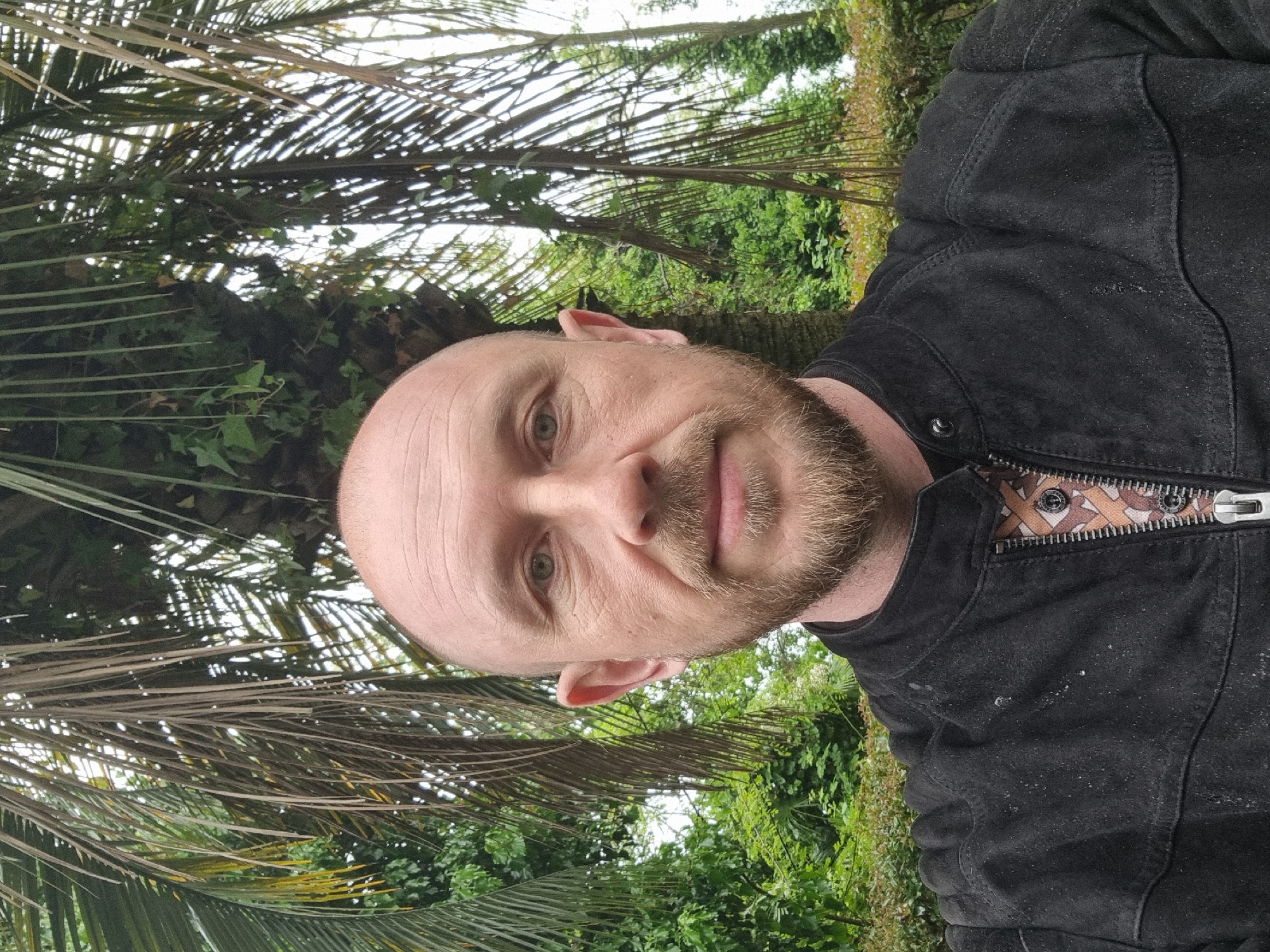 A close-up portrait shows a middle-aged Caucasian man with a shaved head and a short beard. He has light skin and green eyes, and he is looking directly at the camera with a slight smile. He wears a dark-colored jacket with a zipper visible at the collar. The background features lush greenery, including palm trees. Considering the presence of palm trees and the general environment, the location is very likely Sochi, Russia.