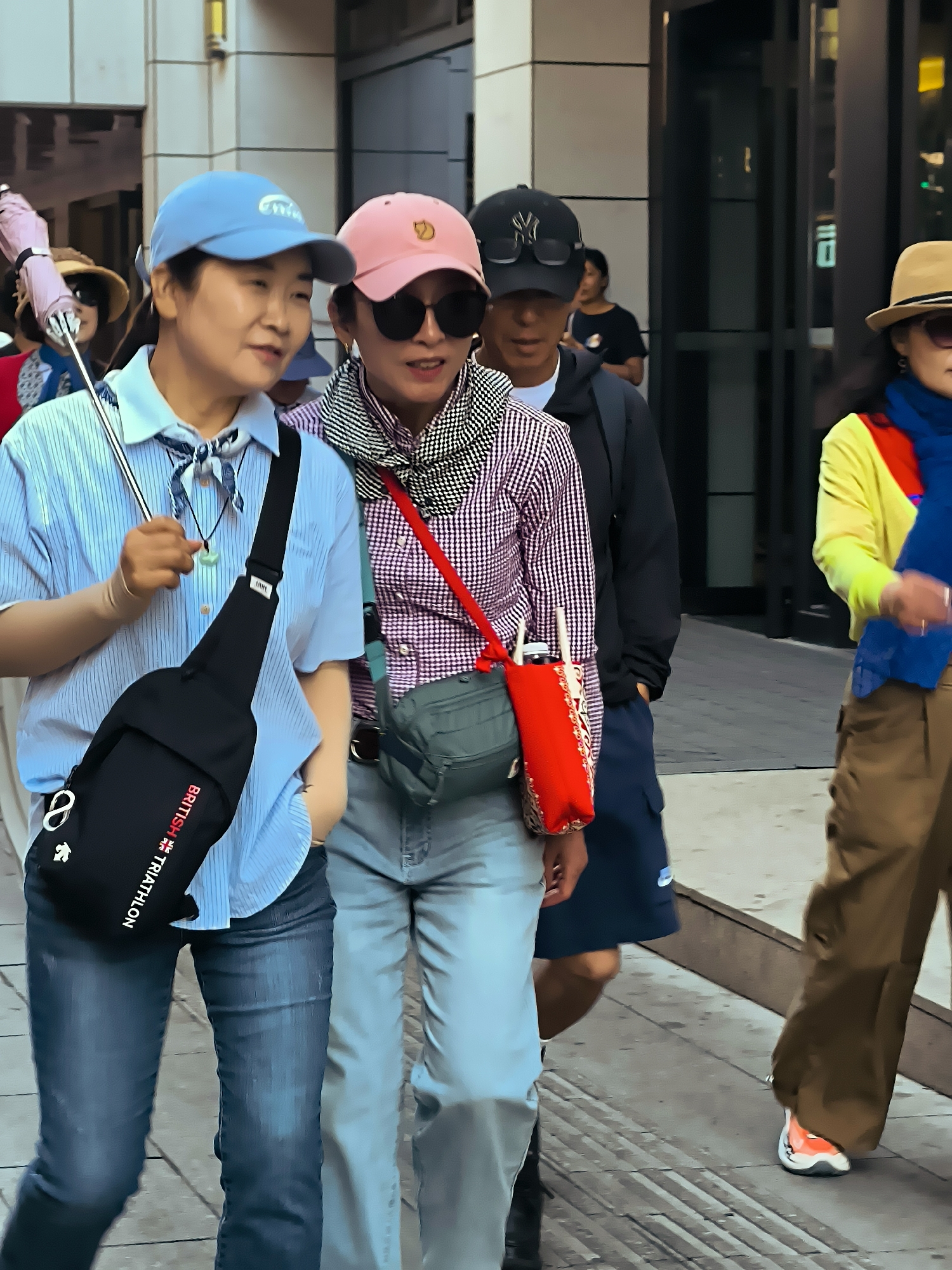 Four individuals are walking on a paved sidewalk in what appears to be a city. The foreground shows two women. The woman on the left is wearing a light blue striped shirt, dark denim jeans, and a light blue baseball cap. She has a black sling bag with red lettering across her chest. The woman on the right is wearing a pink baseball cap, a black and white checkered shirt, and light blue jeans. She is carrying a red bag over her shoulder. Behind them, a person wearing a black baseball cap and sunglasses is visible, along with another individual in the background. To the far right, a person in a tan hat and a brightly colored yellow and red top is walking. The environment suggests an urban setting with buildings and street elements. There is no specific visual information that definitively places the location in Ürümqi, China.