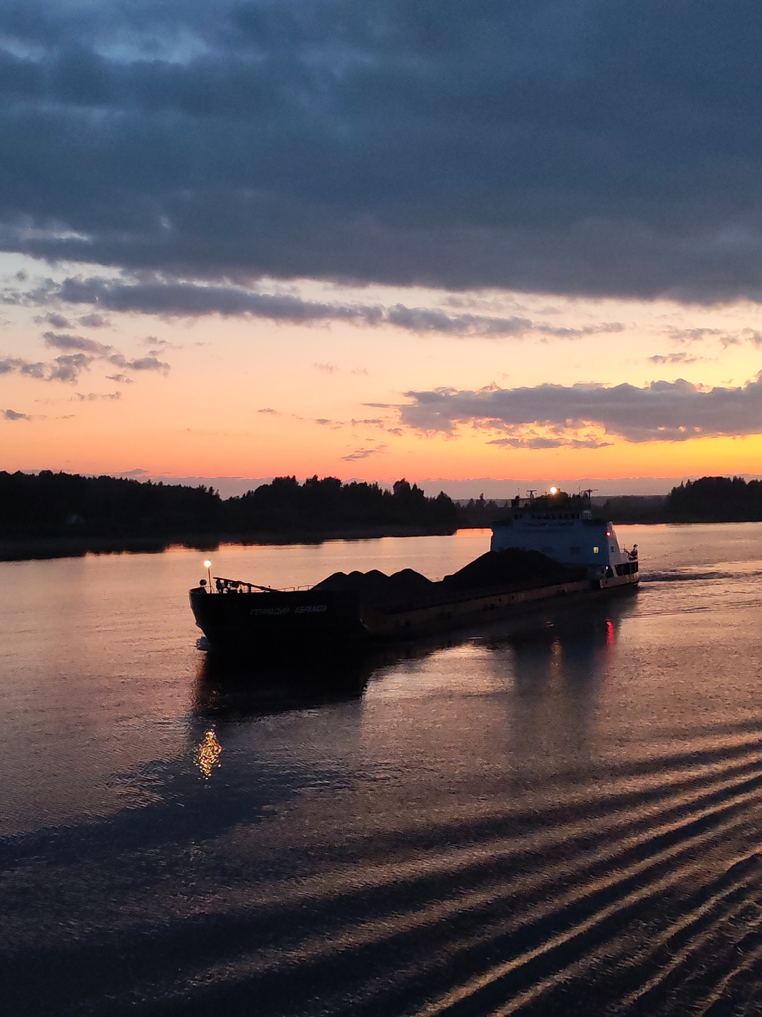 A long barge laden with a dark cargo moves across a body of water, likely a river or wide canal, at dusk. The sky transitions from a dark, clouded blue above to orange and yellow hues near the horizon. Silhouettes of trees line the shore in the background. The water's surface reflects the colors of the sky and the lights of the barge. Wake patterns spread out from the stern of the barge. The location of the image is not mentioned. There are no people visible in the image.