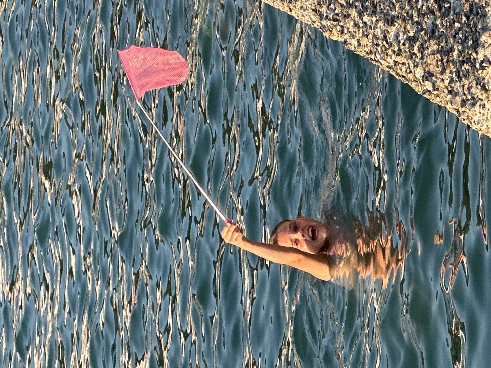 A young boy is swimming in the water near a concrete structure, holding a pink butterfly net. The boy is looking at the camera with his mouth open, appearing excited. The water is a deep blue-green with visible ripples and reflections. The concrete structure is rough and textured, appearing to be a breakwater or pier. The scene takes place in the city of Sochi, Russia, likely during daytime given the bright lighting and reflections on the water.