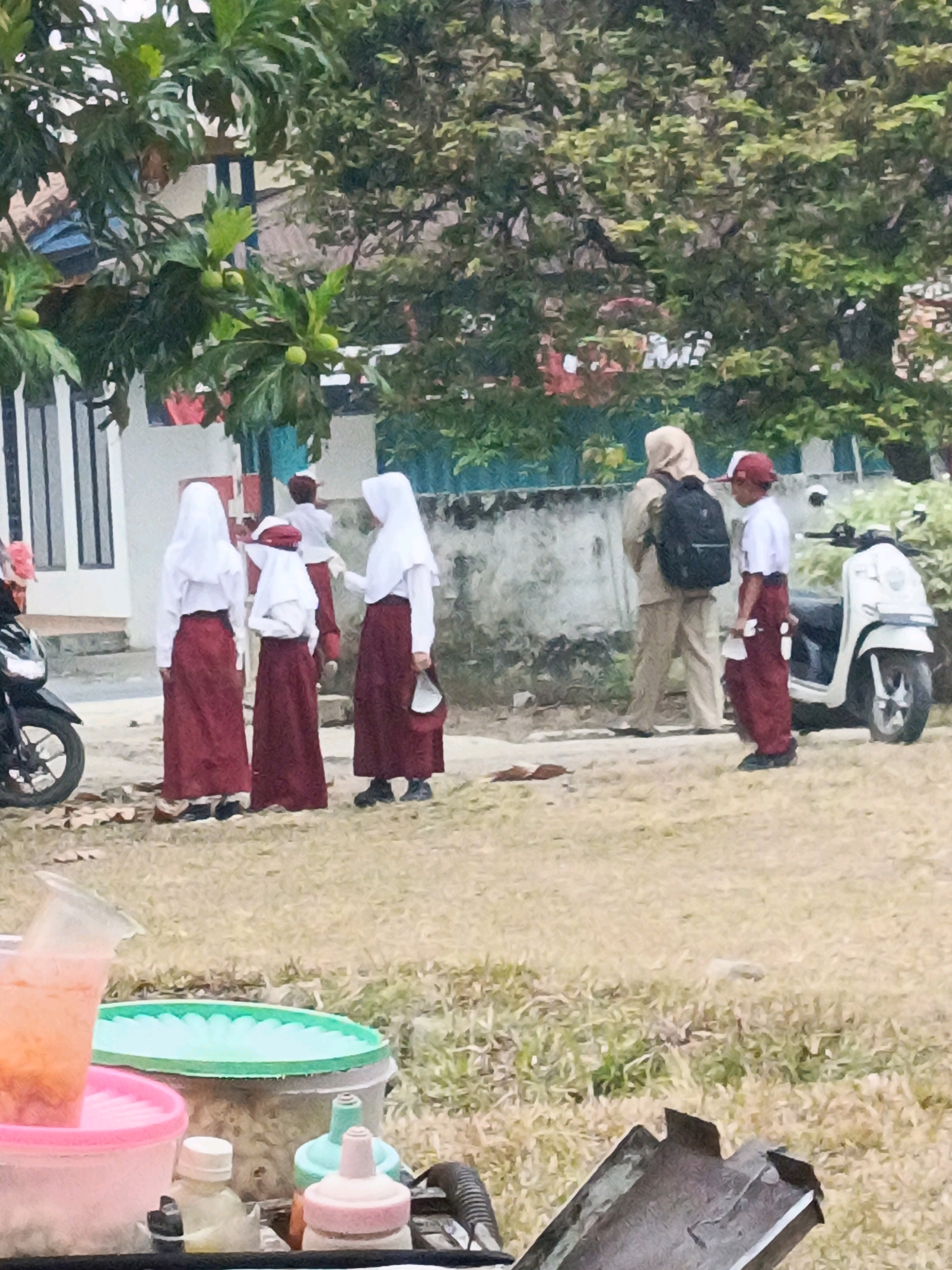 In Megang Sakti II, Indonesia, the scene captures an outdoor setting with several individuals, likely students, gathered near a building and trees. In the foreground, a cart is partially visible with containers of colorful drinks, suggesting a street vendor or a refreshment stand. To the left, a motorcycle is parked.
In the middle ground, a group of four individuals are present, appearing to be students in uniform. Three of them are wearing white shirts and maroon skirts, along with white headscarves. One of these students is holding a white bag or handkerchief. The fourth student is wearing a white shirt and maroon pants with a red hat. Behind them, two adults are also visible. One is wearing a beige uniform and a beige headscarf, carrying a black backpack, while the other is in a white shirt and maroon pants with a red hat, likely another student. A white scooter is parked to the right of the adults.
The background features a building with a white wall and some windows, partially obscured by trees with green foliage and what appear to be green fruits. A low concrete wall runs along the edge of the property. The ground is covered in dry grass. The overall lighting suggests an overcast day.