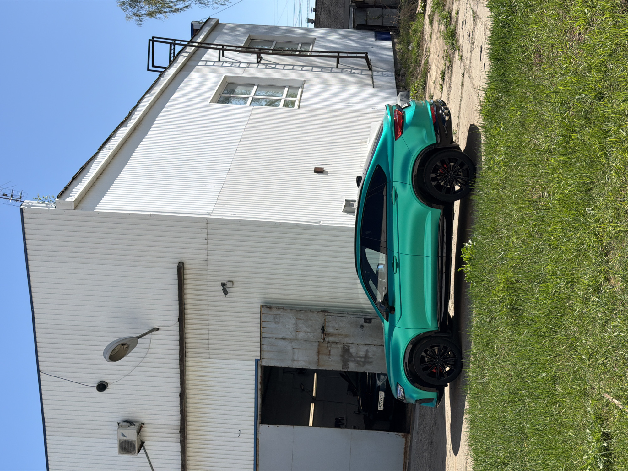 The image depicts a teal-colored car parked in front of a white, corrugated metal building. The building has a large open garage door, and another car is partially visible inside. A street lamp and air conditioning unit are mounted on the side of the building. A black metal ladder runs up the side of the building. The license plate of the car visible inside the garage reads "P577KK 159." The car is parked along the edge of a concrete path, with grass growing alongside. Based on the license plate (159) and some other context clues such as the general building/infrastructure style, the location is most likely Chaikovsky, Russia.