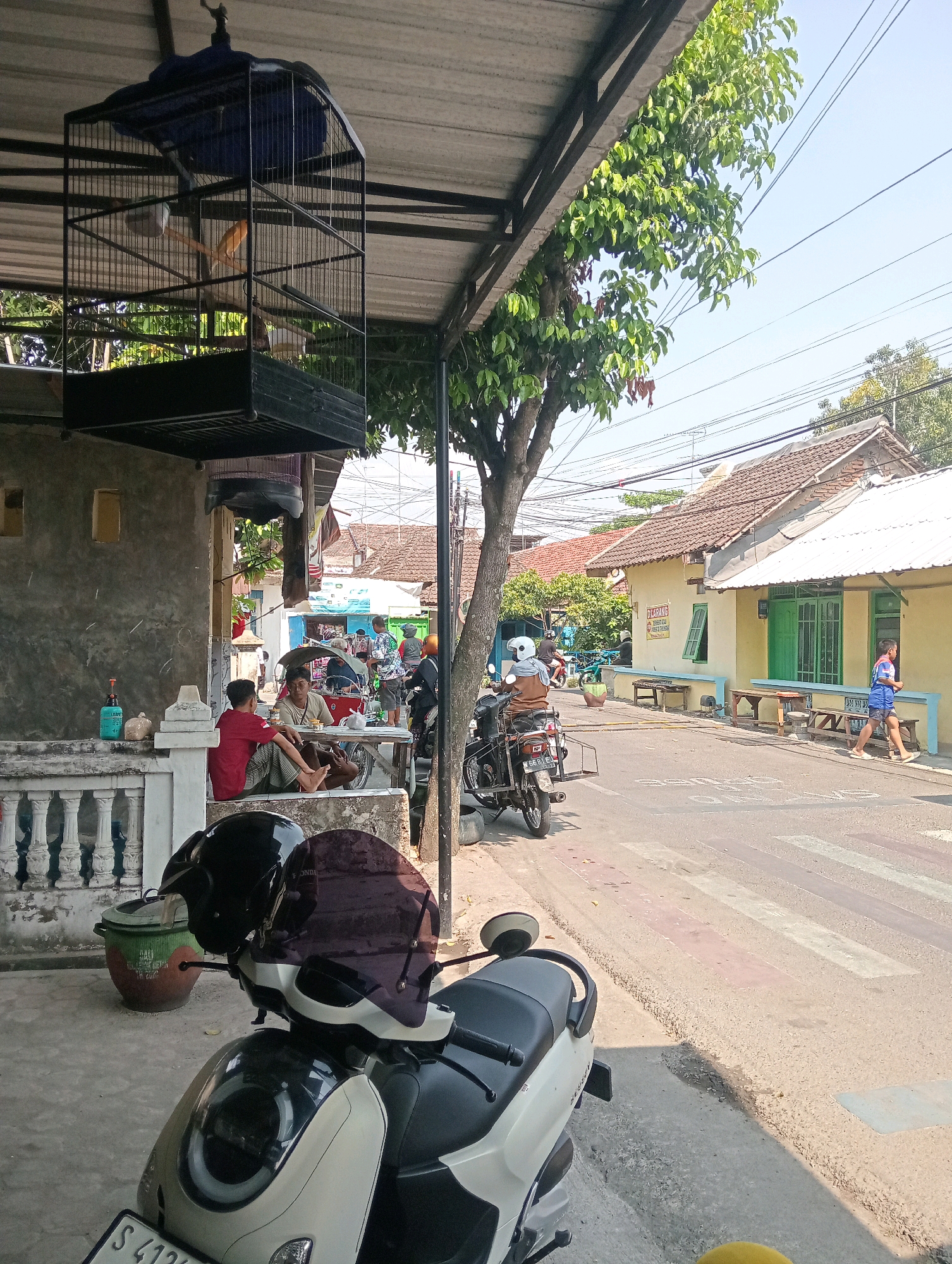 The image depicts a street scene in Bojonegoro, Indonesia, on a sunny day. In the foreground, a white and black scooter is parked, with a black helmet and a dark tinted visor resting on its handlebars. To the left, a large green and brown ceramic pot is visible next to a white balustrade. A birdcage with a bird inside hangs from a metal structure above. Further back, two men are seated at a wooden table on a concrete porch, with one wearing a red shirt and the other a light-colored shirt and sarong. A light blue bottle and some bagged items are on the porch.
The street itself is paved with asphalt and features a pedestrian crossing marked with white and pink stripes. A tree with green leaves grows beside the road, and power lines crisscross the sky. On the right side of the street, several yellow-painted houses with green window frames and white roofs are visible. A few wooden benches are placed outside these houses. People are seen walking and riding motorcycles along the street. One person in a blue shirt and shorts is walking on the sidewalk, and a woman wearing a helmet and a brown top is riding a motorcycle with a sidecar. In the background, other buildings and trees suggest a residential or commercial area within the city. The overall atmosphere is that of a typical day in a bustling Indonesian town.