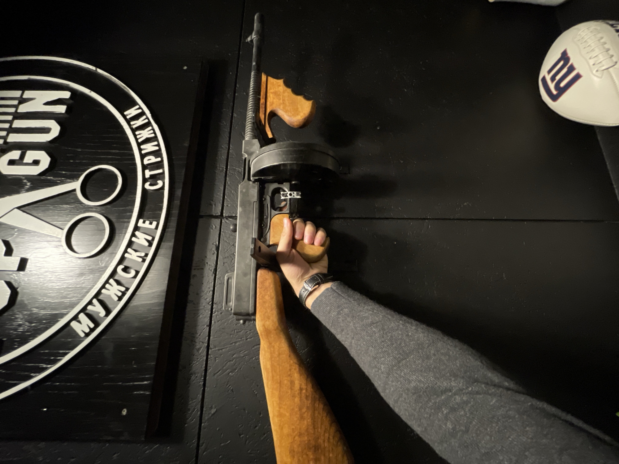 The image shows an interior setting with a black wall. On the left is a circular sign with the word "GUN" vertically stacked, along with scissors and Cyrillic text. A Thompson submachine gun is mounted on the wall. A person's hand is holding the gun. A New York Giants-themed football is on the right side of the wall. The location of the image is inferred to be Saint Petersburg, Russia, based on the Cyrillic text on the sign.