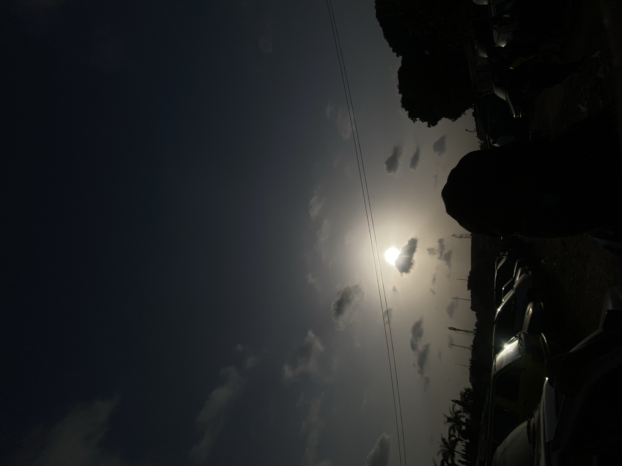 The image, when viewed in correct orientation, depicts an outdoor scene bathed in the dramatic light of either sunrise or sunset, creating strong backlighting and prominent silhouettes. The sky fills most of the frame, transitioning from a dark grey-blue on the left to a bright, hazy white and yellow glow around the sun on the right. The sun, a brilliant circular disc, is positioned in the upper right quadrant, partially veiled by a small cloud which creates a pronounced halo effect. Several other scattered, dark clouds are visible across the sky.
Two thin, dark power lines or cables extend diagonally across the mid-ground of the sky. The lower and right sections of the image are dominated by heavily silhouetted elements. These include dense, dark foliage, with distinct palm trees visible towards the bottom right. A row of vehicles, likely cars or trucks, appears parked or lined up along the lower right, their forms mostly obscured by shadow, though some reflective surfaces catch glints of light. A large, dark silhouette, possibly a person's head and shoulders or a substantial object, is prominently situated on the far right, partially obscuring the view of the vehicles. In the distance, faint outlines of structures or poles can be discerned between the vehicles and the horizon. The strong contrast and backlit subjects indicate a time of day when the sun is low in the sky. No text is visible in the image.