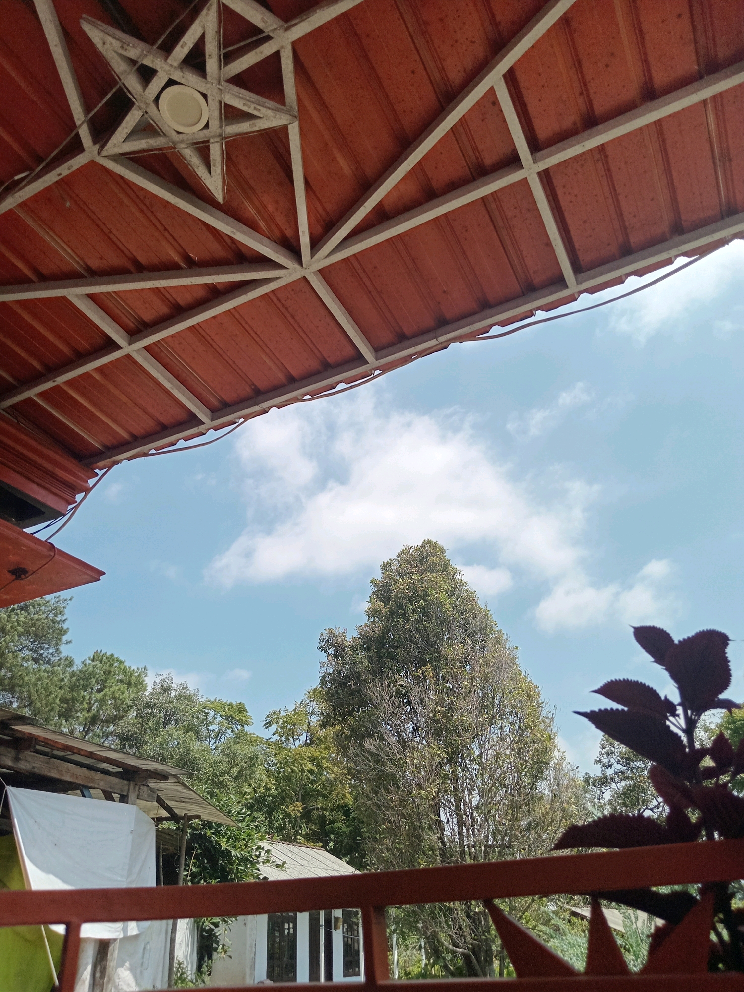 A low-angle, medium shot captures a view of the sky framed by a reddish-brown corrugated metal roof supported by a white metal frame. In the center of the frame, a decorative white star-shaped element is visible. Below the roof, a cluster of trees and vegetation fills the middle ground. To the left, a rustic structure with a white tarp is partially visible. In the foreground, a reddish-brown railing with decorative pointed elements runs across the bottom of the image. The sky is a light blue with scattered white clouds. The scene appears to be an outdoor area, possibly a patio or veranda, in a natural setting.