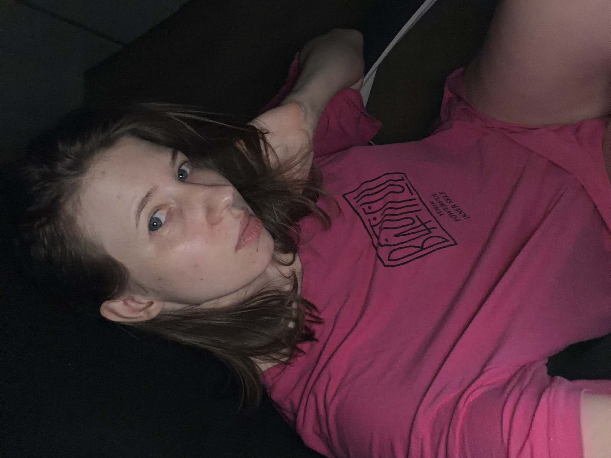 A young woman with light skin and blue eyes is pictured from the chest up. She has medium-length brown hair that falls around her shoulders and is wearing a bright pink t-shirt with a black graphic design on it. The graphic design appears to be text, with the words "YOUR POWERFUL INNER SELF" visible below a larger, stylized word. Her left arm is bent, and her hand is resting near her head. The background is dark and appears to be indoors, with some shadows suggesting the presence of furniture. The lighting is dim, with some ambient light illuminating the woman's face and upper body. There is no visible indication of the city of Dongducheon-si, South Korea.