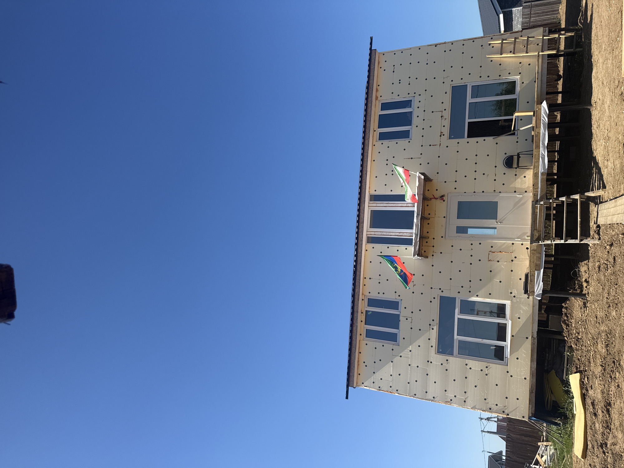 A two-story, unfinished house is shown under a clear blue sky in the city of Matchino, Russia. The house is covered in a light-colored insulating material with black circular fasteners. Two flags, one with green, white, and red stripes, and another with green, white, red, and blue stripes, are attached to the front of the house. The house has several windows and a white door with a small overhang. A wooden deck with stairs leads up to the entrance, and a ladder leans against the side of the house. Construction materials, including yellow foam insulation, are visible on the ground. A chair and a piece of wood are on the deck. The surrounding area is a dirt construction site with a wooden fence in the background.
