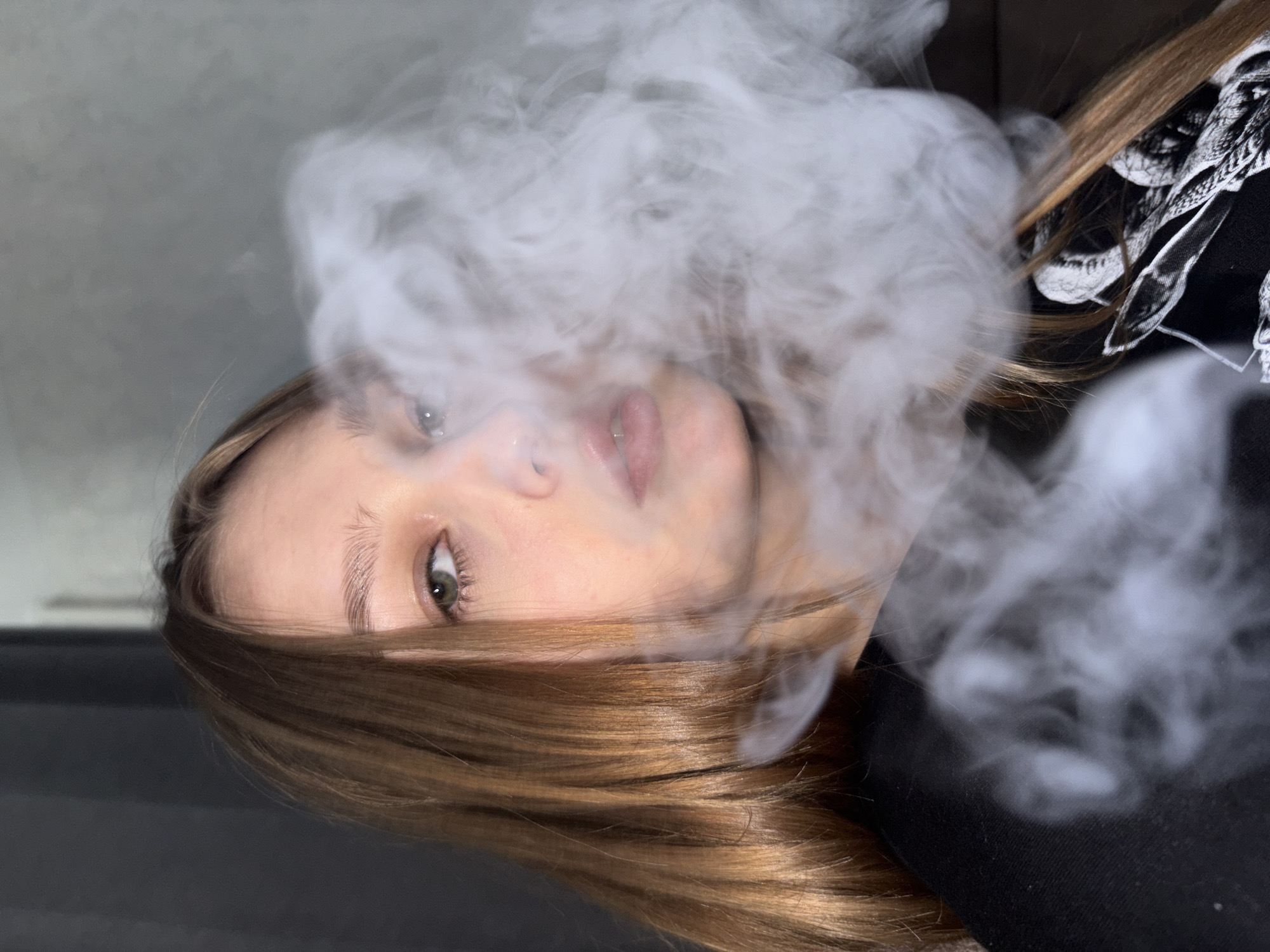 A close-up, angled shot of a woman's face and upper body obscured by a cloud of vapor. Her light brown hair is visible framing her face, with some strands partially covering her left eye. She has fair skin, with visible eyebrows and eyelashes. Her eyes are partially visible, and her lips are slightly parted. She is wearing a black shirt or top, with what appears to be a white patterned design. The background is a gray-black color, which appears to be a wall or surface. No identifiable location in the city of Марковский, Russia can be determined from the image. The primary action is the exhalation of vapor, covering a significant portion of the subject's face.