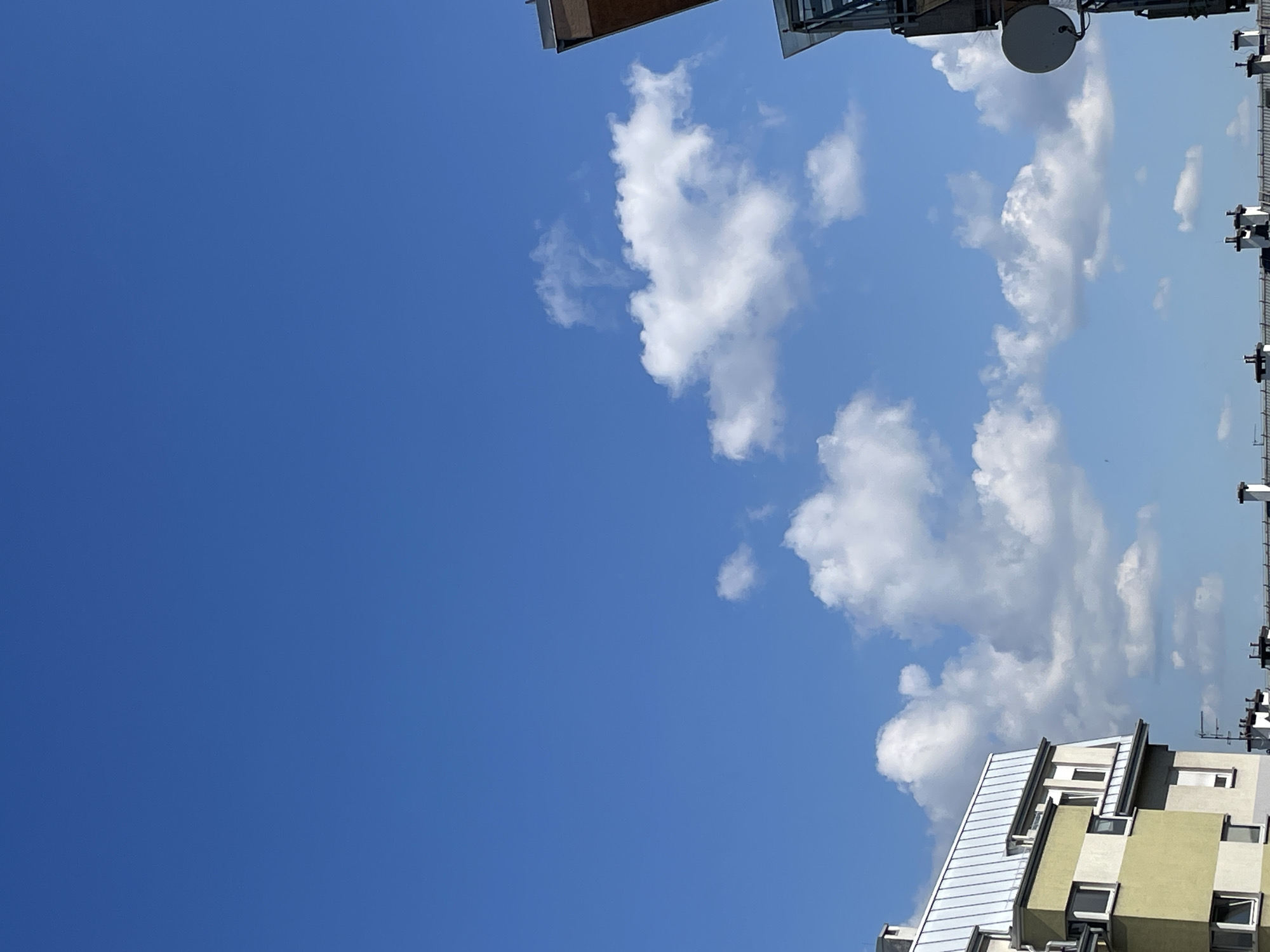 The image is an upward-angled shot of a clear, bright blue sky dotted with scattered white cumulus clouds. In the bottom right and top right corners of the frame, parts of buildings are visible, including rooftops, satellite dishes, antennas, and exterior walls in shades of tan, grey, and white. The buildings' architectural styles appear modern. The image is taken from the location of Unknown, Unknown.