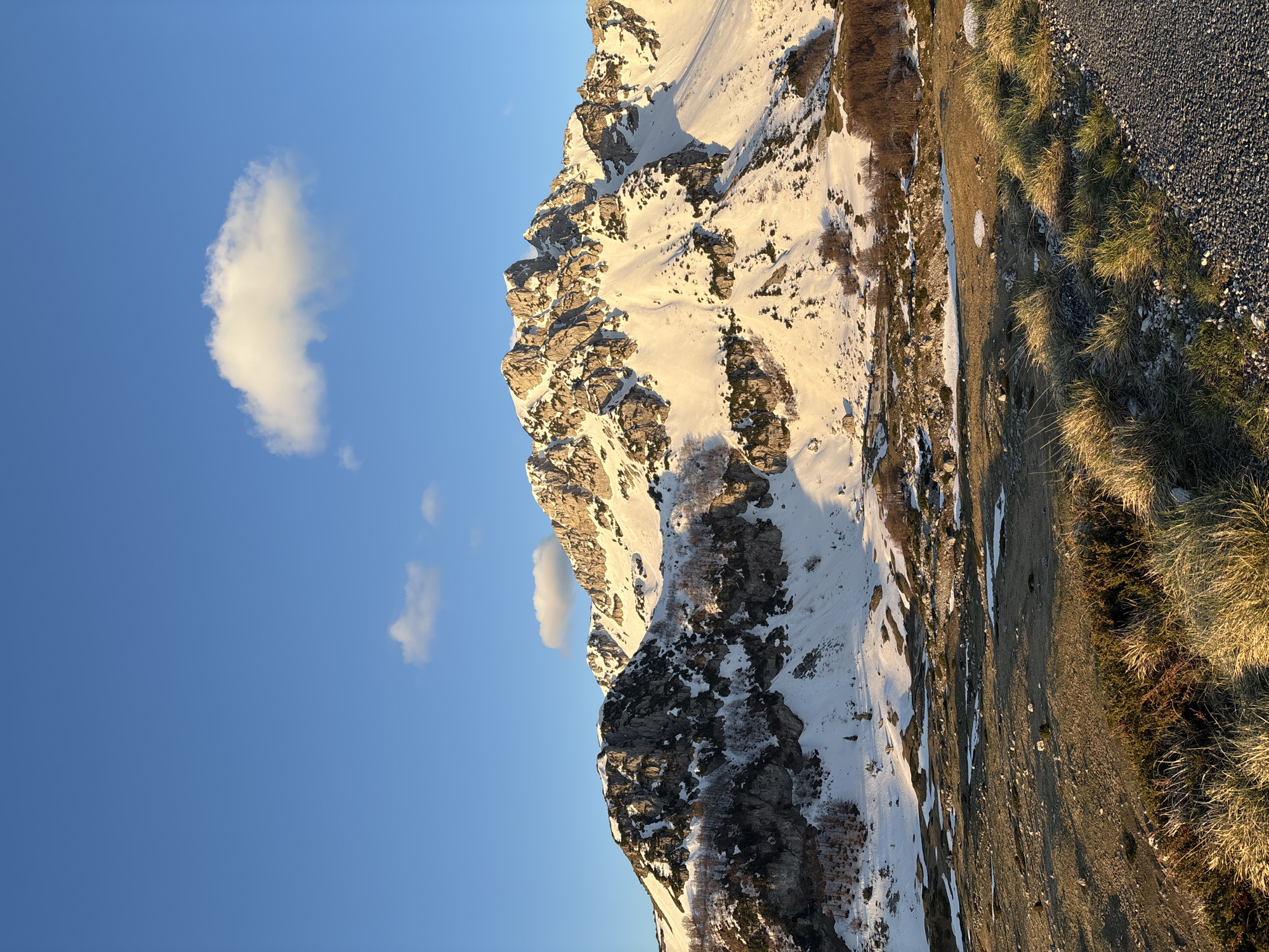 The image shows a mountain partially covered in snow in Pašina voda, Montenegro. The left side of the image has a blue sky with a few small, white clouds. The right side shows a grassy area and what appears to be a gravel road. The mountain dominates the center of the frame, with snow clinging to its rocky faces and slopes. The sun appears to be low in the sky, casting shadows across the scene.