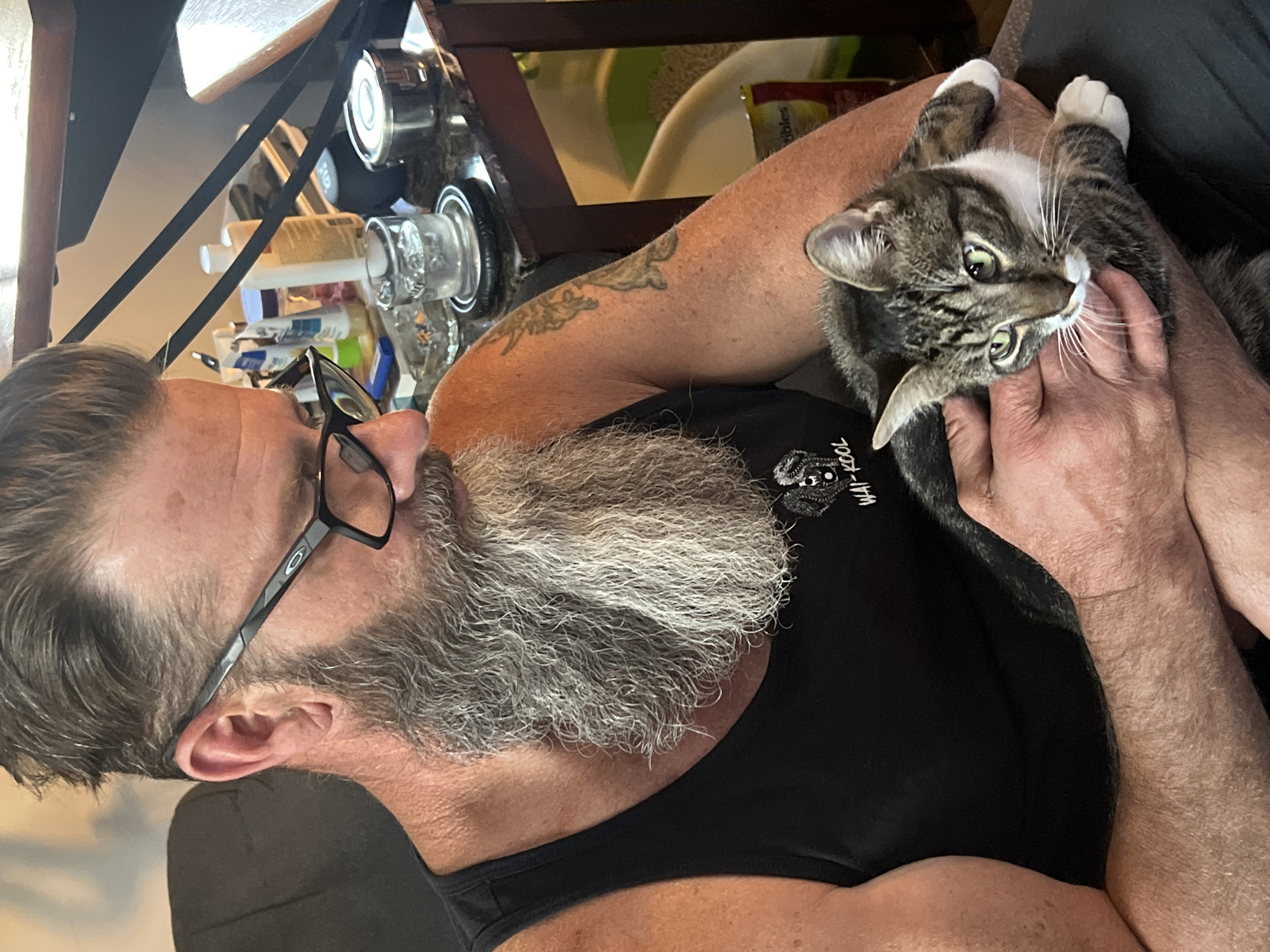 A close-up, candid shot shows a man with a graying beard and glasses holding a tabby cat with white paws. The man is wearing a black tank top. A table cluttered with miscellaneous items sits in the background. The man appears to be in a residential indoor location. There is no information to confirm the location is the City of Newburgh, United States of America.