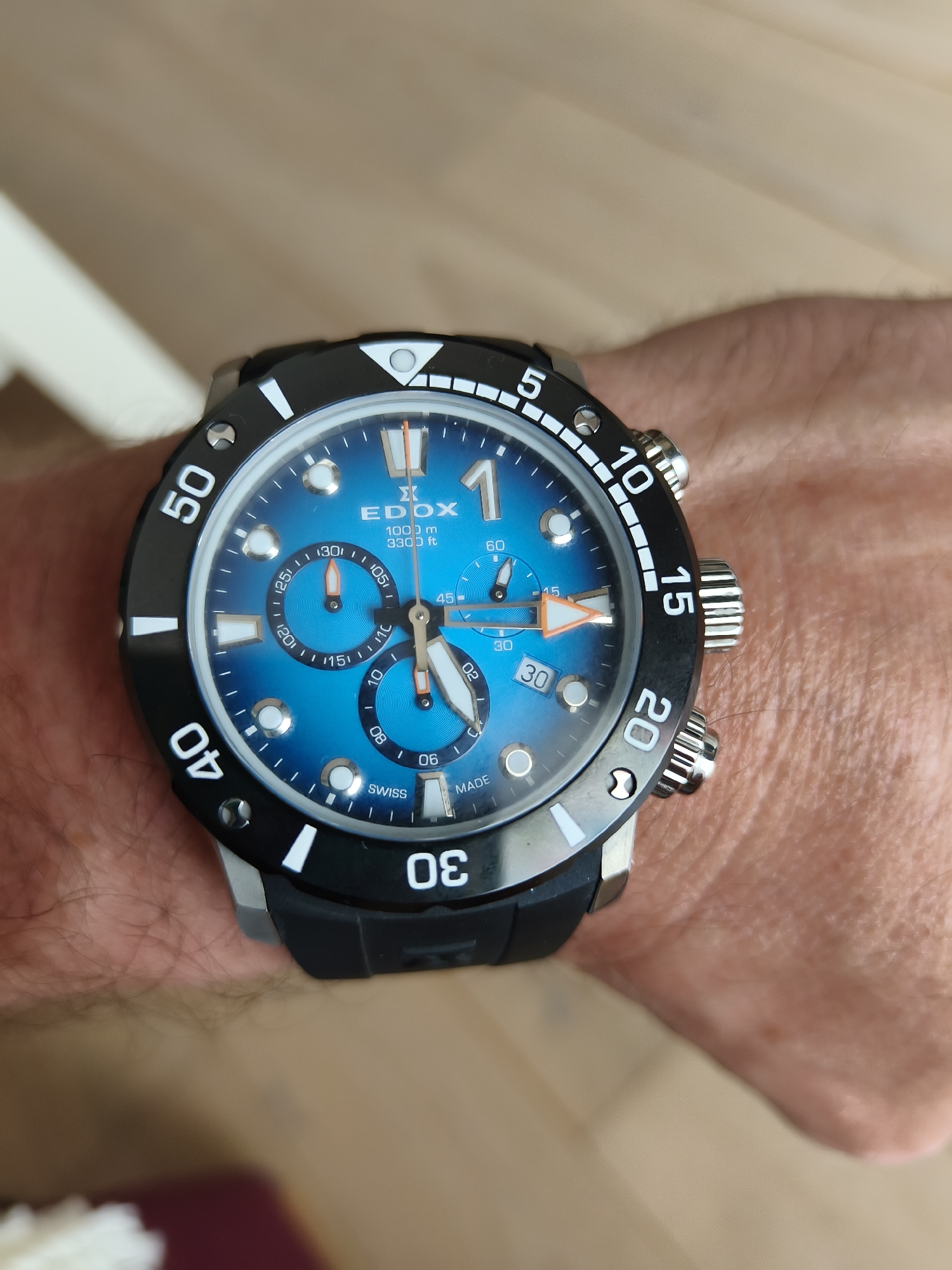 The image shows a close-up of a person's wrist wearing an Edox watch with a blue face and black bezel. The watch features chronograph dials and is marked with "1000 m 3300 ft" indicating its depth rating. The background is slightly blurred, revealing the person's skin and a glimpse of a light-colored surface, possibly a table or floor.