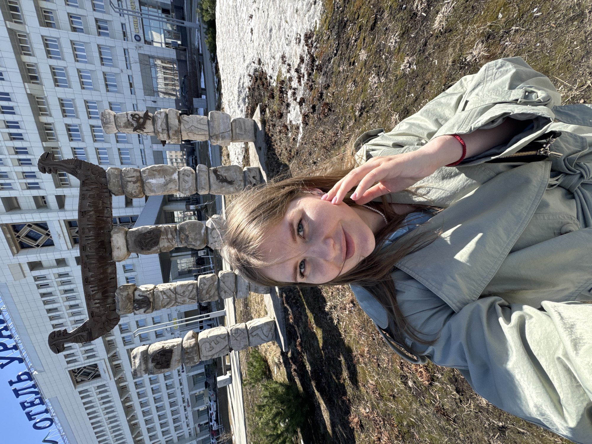 A young woman takes a selfie outside in Perm, Russia. The photograph captures the subject from a tilted, close-up perspective, showing her face, hair, and upper body. She is wearing a light green jacket and has her hand near her face.
Behind the woman is a sculptural monument constructed of carved stone blocks, crowned by a dark, metal piece resembling an animal head or tribal figure. Adjacent to the monument is a large, multi-story building with repetitive window patterns, typical of Soviet-era architecture. In the background, residual patches of snow are visible on the ground. The ground around the woman appears to be covered in dry grass and fallen leaves. A blue banner with Cyrillic text is visible in the lower left corner of the image.