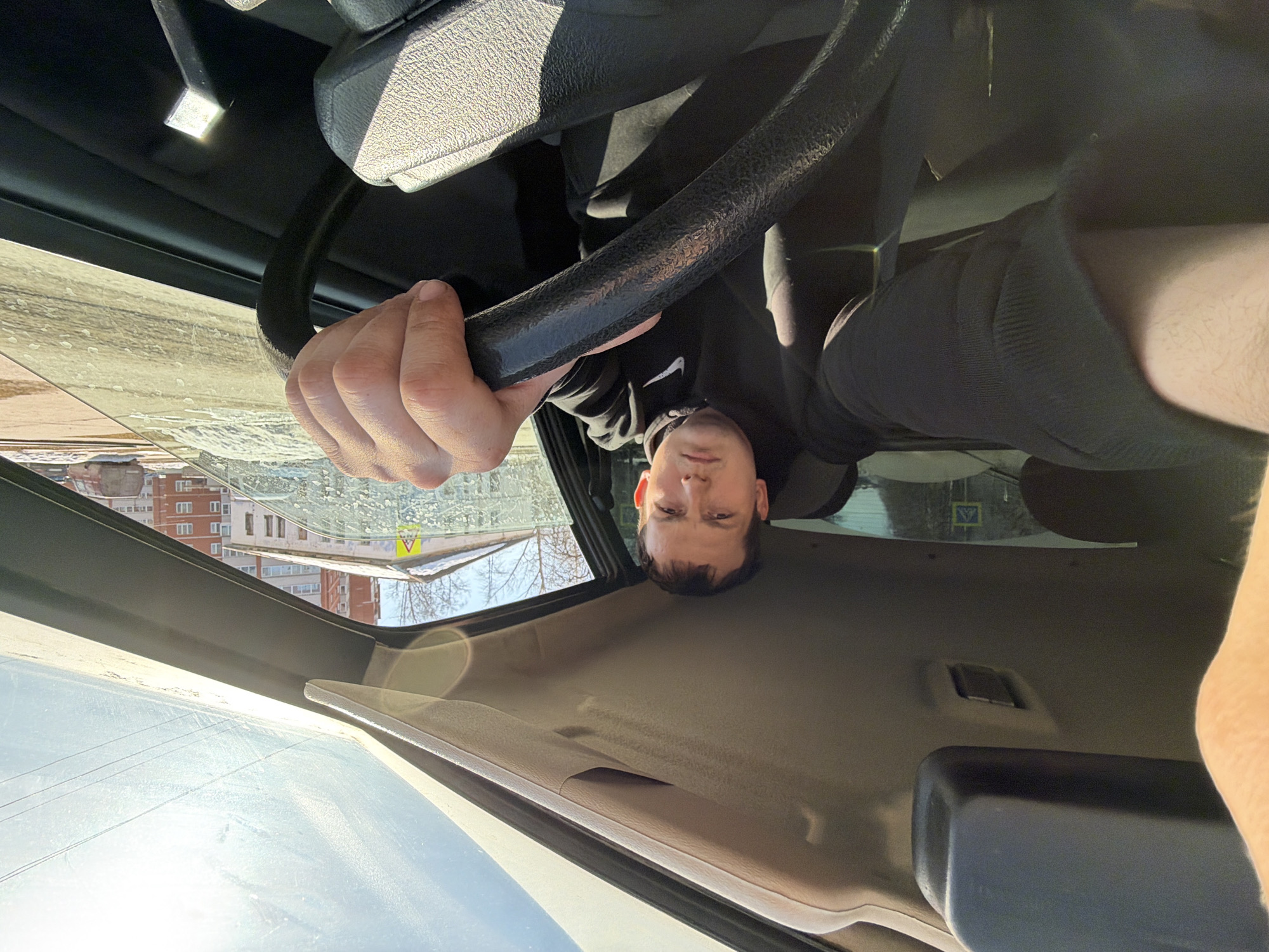 A man is inside a car holding the steering wheel. Outside the car is a building and a street sign in Russian indicating the location is likely in the city of Chaikovsky, Russia.