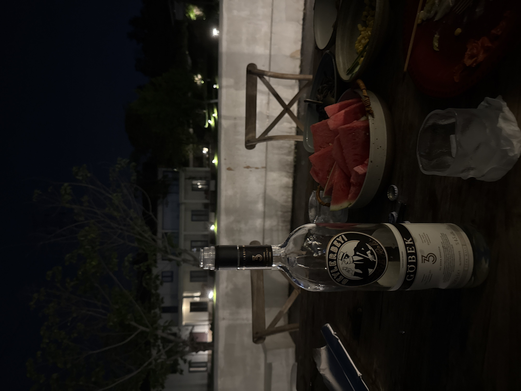 The image depicts a table setting at night. A bottle of Beylerbeyi Göbek rakı lies on the table amidst plates of food, a bowl of sliced watermelon, a glass with ice, and miscellaneous objects. In the background, lights suggest a building or street scene at night. The location is not identifiable as Jinhua, China, from the information provided.