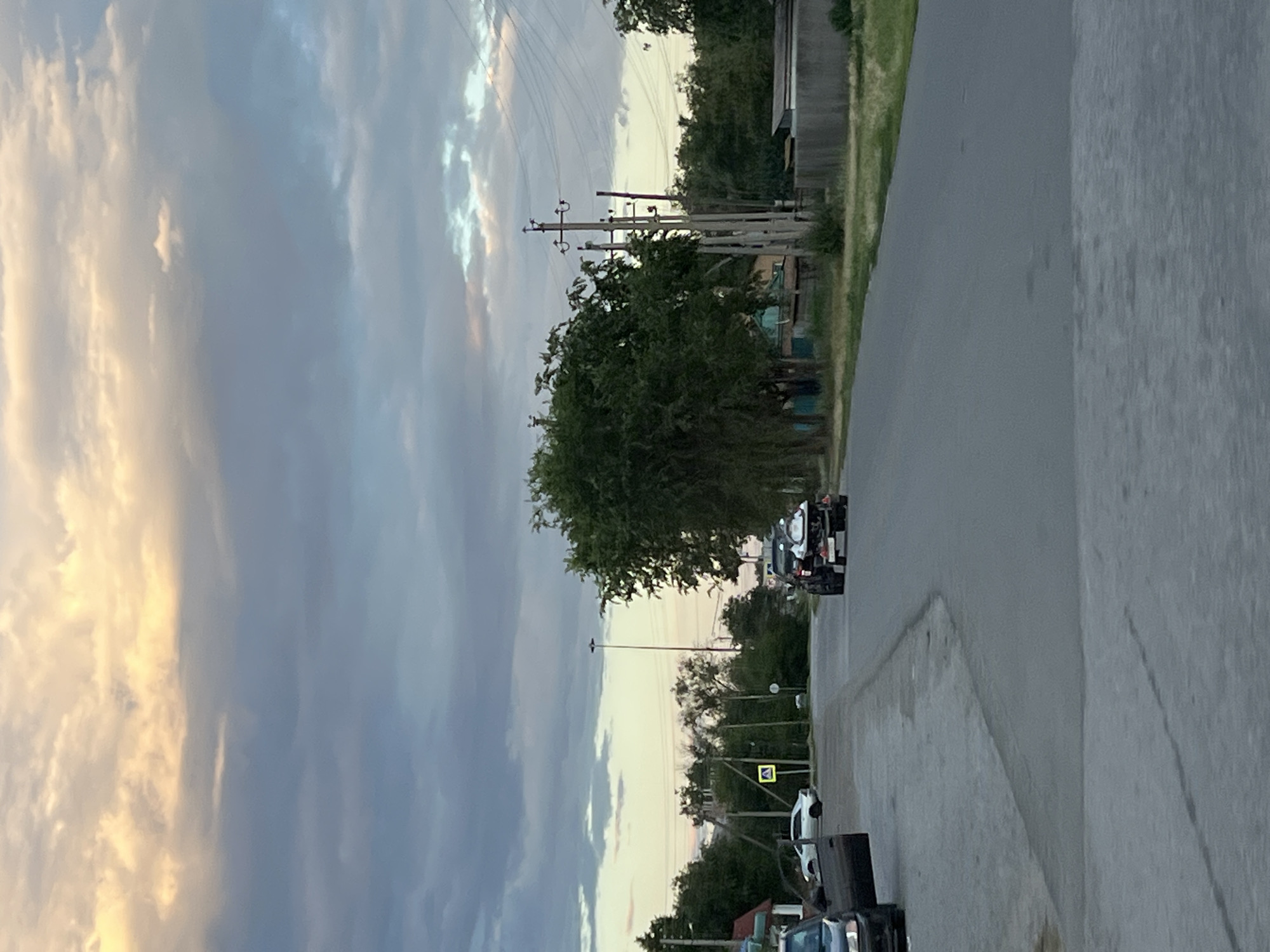 The image depicts a roadside scene in Zimovniki, Russia, likely during the late afternoon or early evening, as indicated by the soft, diffused light illuminating the sky. The sky is a dramatic canvas of clouds, with streaks of golden light breaking through the predominantly gray and blue tones, suggesting a transition from day to night or a clearing after rain.
The foreground shows a gray asphalt road curving into the distance. On the right side of the road, there is a concrete embankment. A few vehicles are parked along the side of the road. One is a white car, and behind it, a dark object, possibly a dumpster or a large piece of equipment, is visible. Further down the road, a black and white truck is parked.
On the left side of the road, a line of utility poles with wires stretches into the distance, a common sight in rural or suburban areas. Lush green trees and bushes line the road, creating a natural barrier between the road and what appears to be a building or structure partially visible in the background on the far left. The trees are dense and full, suggesting a temperate climate and a time of year with active vegetation growth. A yellow pedestrian crossing sign is also visible on a utility pole.
The overall atmosphere is one of quietude and everyday life, with no people overtly visible in the scene. The natural elements, particularly the sky and the vegetation, are prominent, creating a sense of place that is both tranquil and slightly dramatic due to the cloud formations. The image is oriented vertically, giving a strong sense of perspective along the road and up towards the sky.