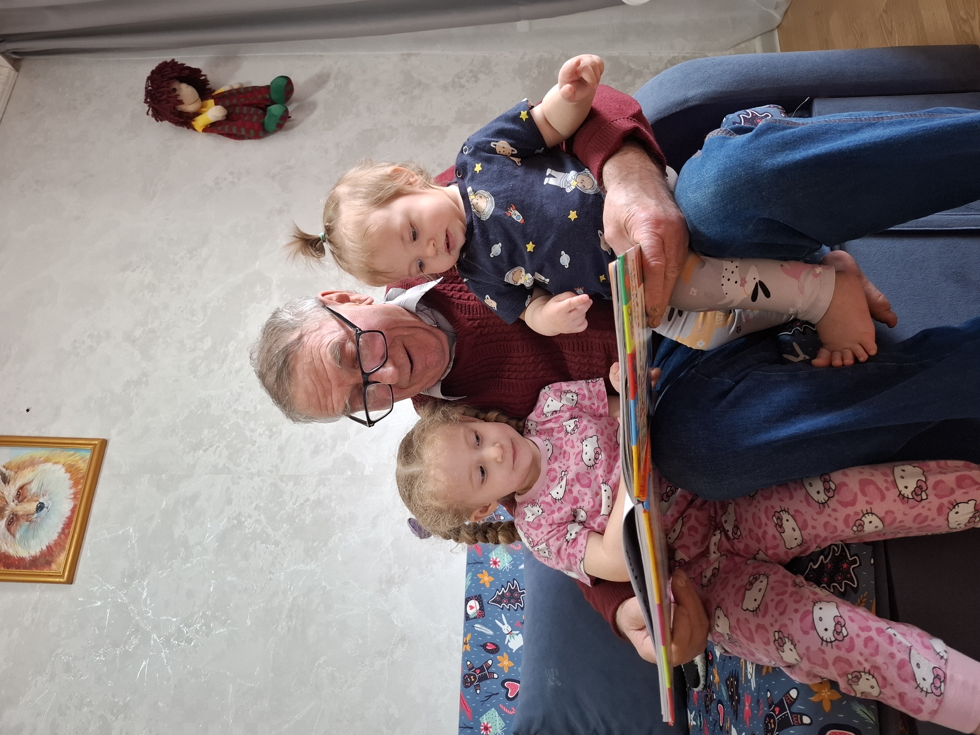 In an indoor setting in Chaikovsky, Russia, an elderly man with glasses sits on a blue cushion with two young children. The man wears a red sweater and blue jeans, and is holding a book. One child is dressed in a navy blue shirt with an astronaut and space design and light-colored patterned leggings, and the other wears pink pajamas with Hello Kitty designs. The walls are painted white with a textured design. A framed picture of a fox and a stuffed doll are visible on the wall.
