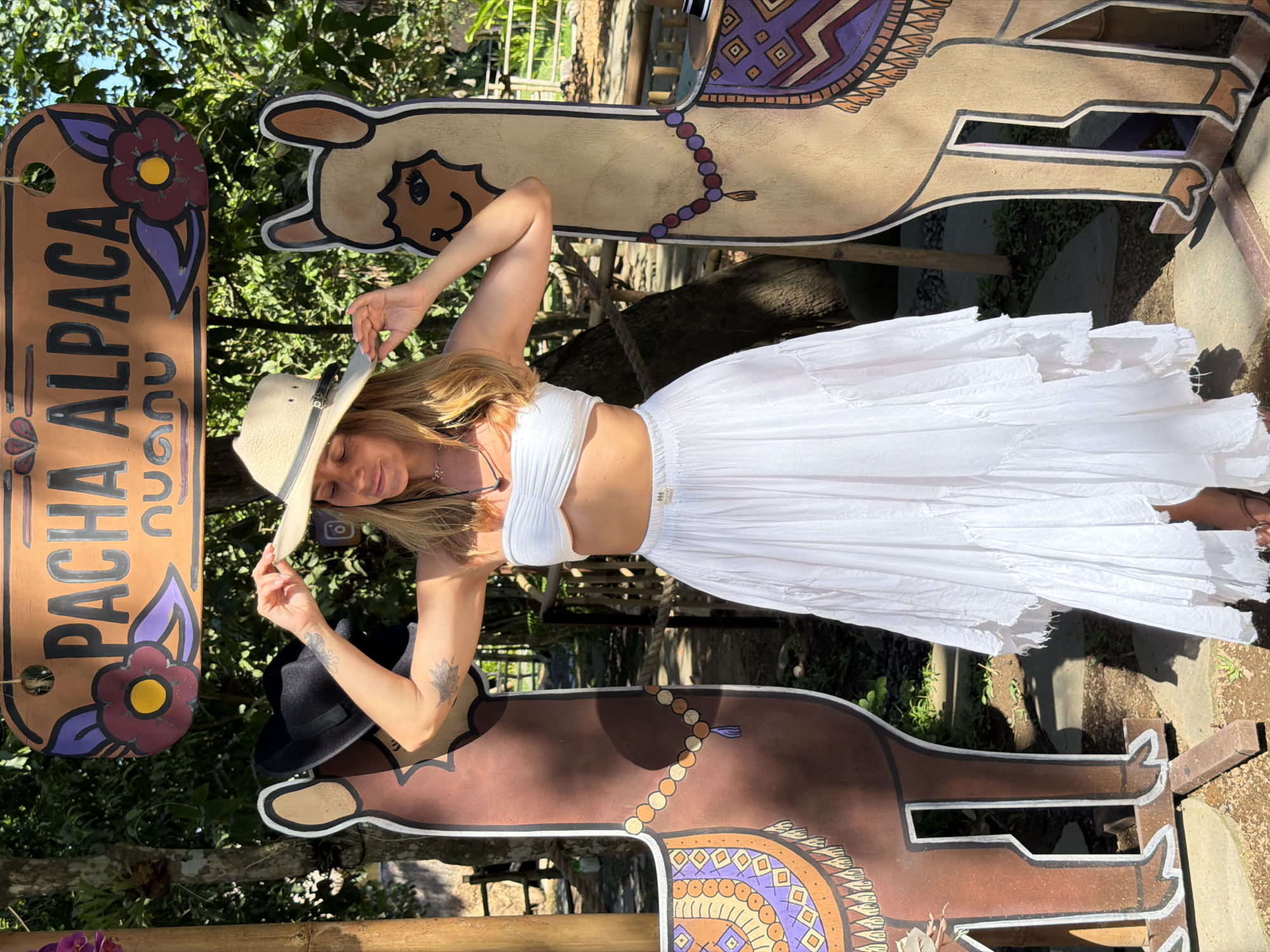 A woman with blonde hair, wearing a white strapless top and a long white flowing skirt, stands outdoors. She is holding a straw hat with a black band, adjusting it on her head. The woman has tattoos on her arm. She is positioned between two cardboard cutouts of alpacas, one on her left that is brown with an orange and purple patterned saddle, and one on her right that is tan with a purple and orange patterned saddle. Above the alpacas, a wooden sign with painted flowers and the words "PACHA ALPACA" is visible. The background consists of lush green foliage and trees, suggesting an outdoor, possibly tropical or subtropical, setting. The ground is covered with dirt and scattered stones. The lighting indicates it is daytime, with sunlight casting shadows. This scene is located in the city of Kediri, Indonesia.