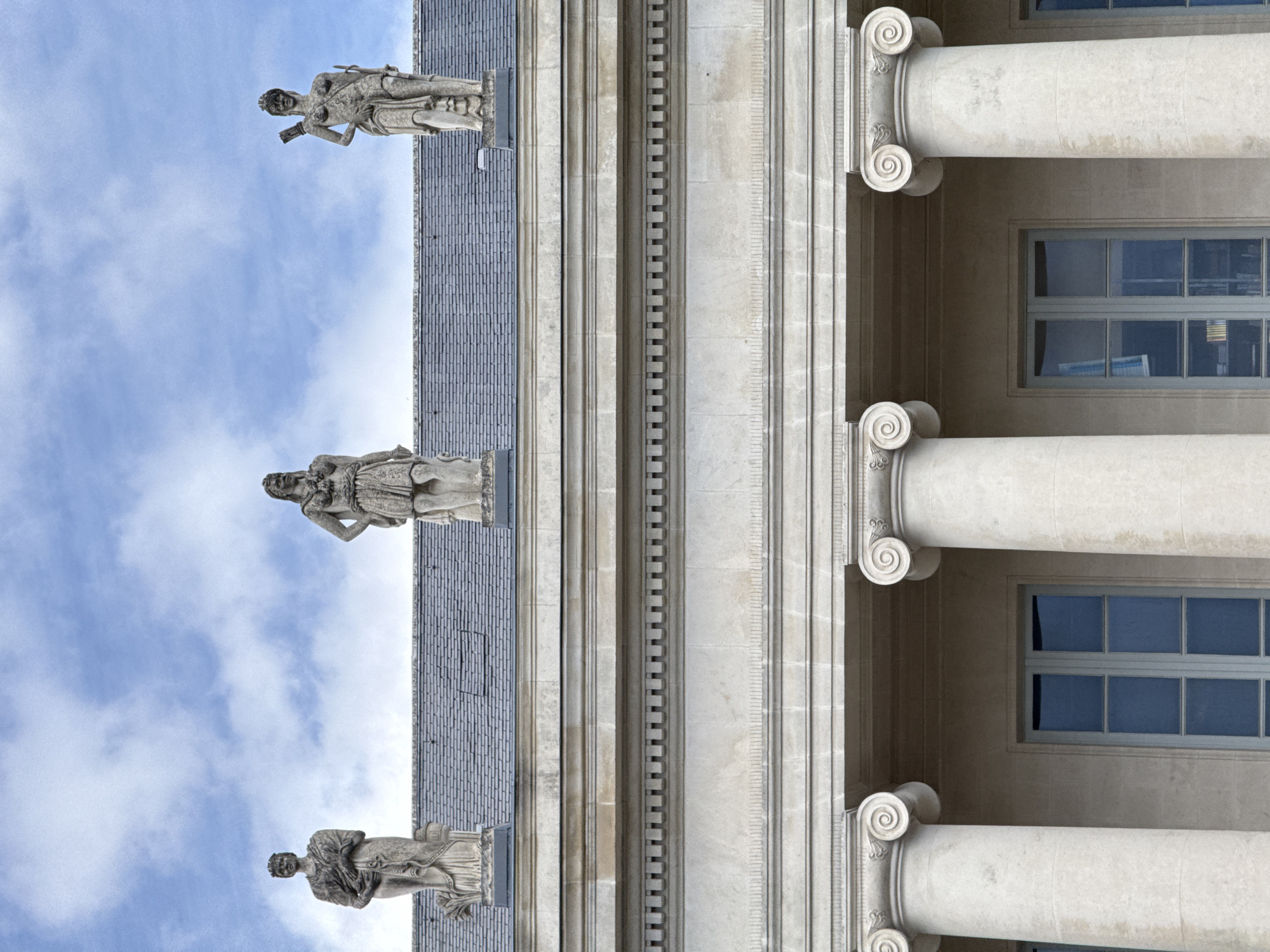 The image shows the exterior of a building with classical architecture. Three stone statues of robed figures stand atop the building's roof. The building features large columns with scrolled capitals and rectangular windows. The sky behind the building is blue with scattered clouds. This is the facade of the Nantes Museum of Fine Arts, located at Place du Lieutenant Colonel Sébert, 44000 Nantes, France.