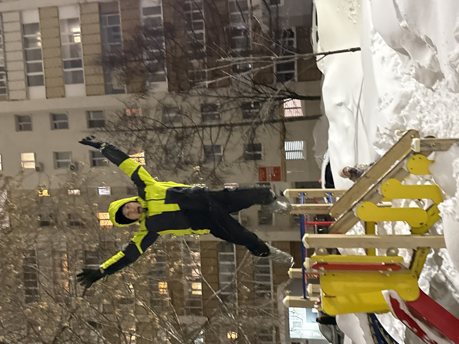 The image captures an outdoor, snow-covered residential area at night in Kazan, Russia. In the foreground, a male individual in mid-air is the central subject, dressed in a black and bright yellow hooded winter jacket, black pants, black gloves, and light-colored boots. His arms are raised, and his mouth is open. He is positioned above a wooden and plastic playground structure, which is partially submerged in deep snowdrifts. The playground features yellow climbing elements and a red slide. In the mid-ground, a second individual, wearing a light-colored beanie and a face covering, is partially visible behind the playground equipment and snow, observing. The background features a multi-story, light-colored apartment building with numerous illuminated windows, indicating nighttime. Several bare deciduous trees stand between the playground and the building. A dark-colored vehicle, partially covered by snow, is visible on the left edge of the frame. The entire scene is under artificial illumination, casting soft shadows.