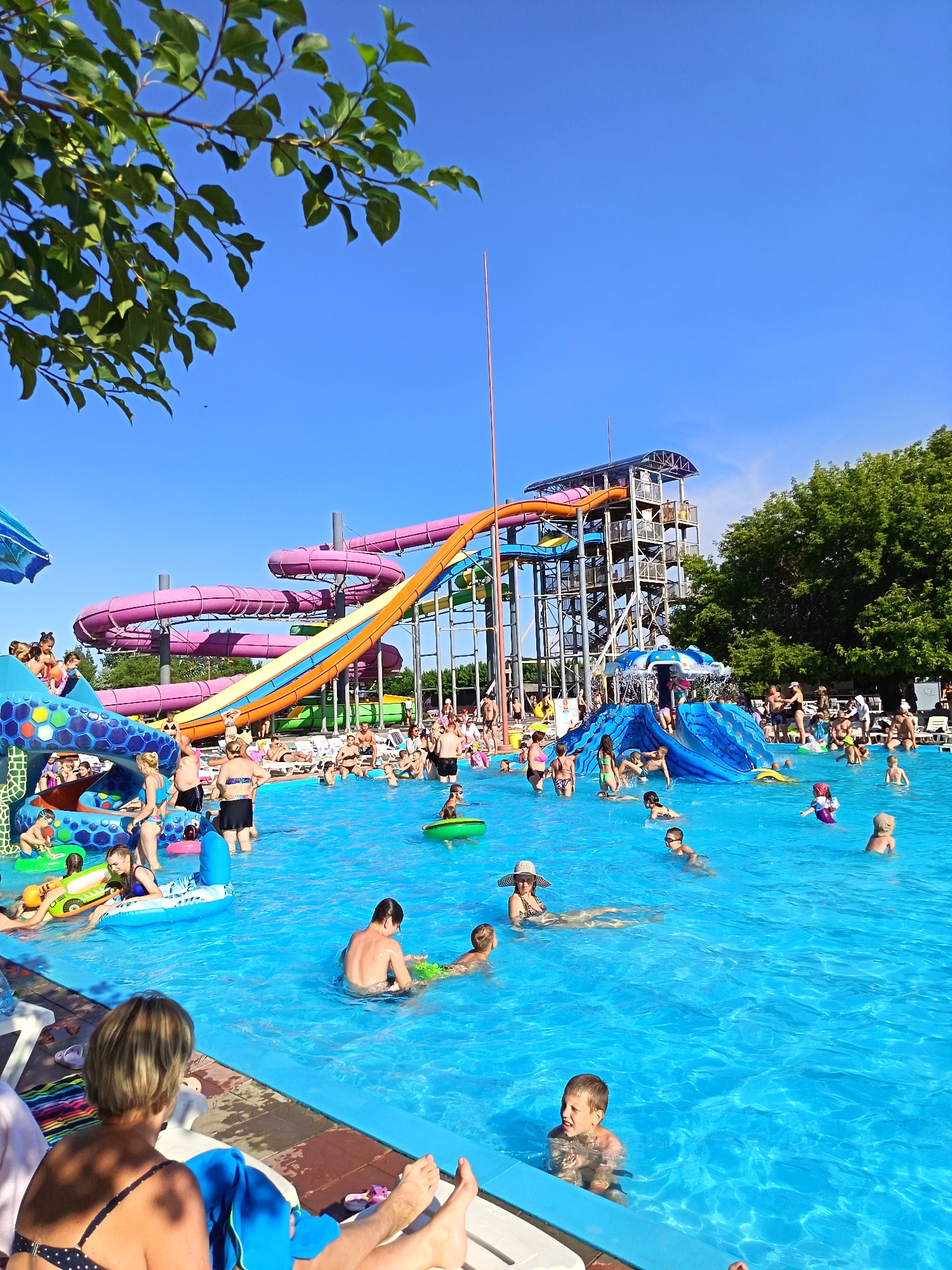 The image depicts a crowded water park on a sunny day, with a large blue swimming pool in the foreground. Several water slides are visible, including large purple spiral slides and a prominent orange and blue slide. People of various ages are enjoying the water, with some swimming, others sitting on inflatable toys, and children playing. There are also lounge chairs and umbrellas around the pool. Trees and a clear blue sky form the background. The location is specified as the water park in Yarovoye, Russia.
