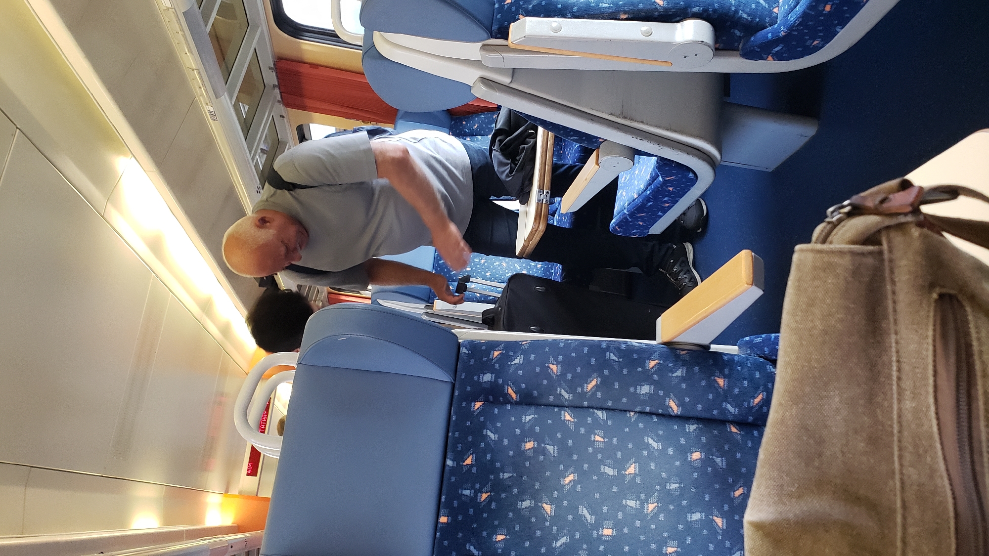 An interior shot of a train carriage in Margecany, Slovakia captures a bald, fair-skinned man, appearing to be in his late 50s or 60s, wearing a grey t-shirt and dark pants. He is standing and looking down, with his right hand on the handle of a black rolling suitcase. Another person, with dark, curly hair, is partially visible behind him. The train seats are upholstered in a blue fabric with a repeating pattern of orange and black geometric shapes. A small fold-down table is extended in front of the man, with a dark fabric item resting on it. A tan canvas bag is prominently displayed in the foreground, partially obscuring the seats. The lighting in the carriage appears to be overhead fluorescent fixtures, with orange accents on the ceiling and walls. Red curtains are visible in the background. The overall scene suggests a travel environment on a train.