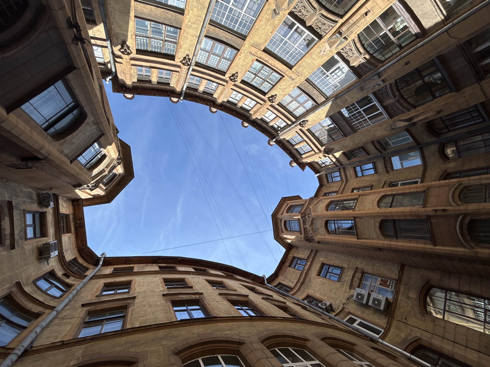 An eye-level, wide-angle, outdoor shot captures the internal courtyard of a multi-story building, viewed from below and looking upwards towards the sky. The architecture is predominantly yellow brick with brown detailing around windows and corners. The buildings curve inwards, creating a sense of enclosure, with arched window frames visible on some levels. The sky is a clear, bright blue with thin white clouds and several dark electrical wires crisscrossing overhead. The image is taken during the day, with sunlight casting shadows on the building facades. There are no people or animals visible in the frame. The scene suggests an urban environment with a distinctive architectural style, possibly from the late 19th or early 20th century.
