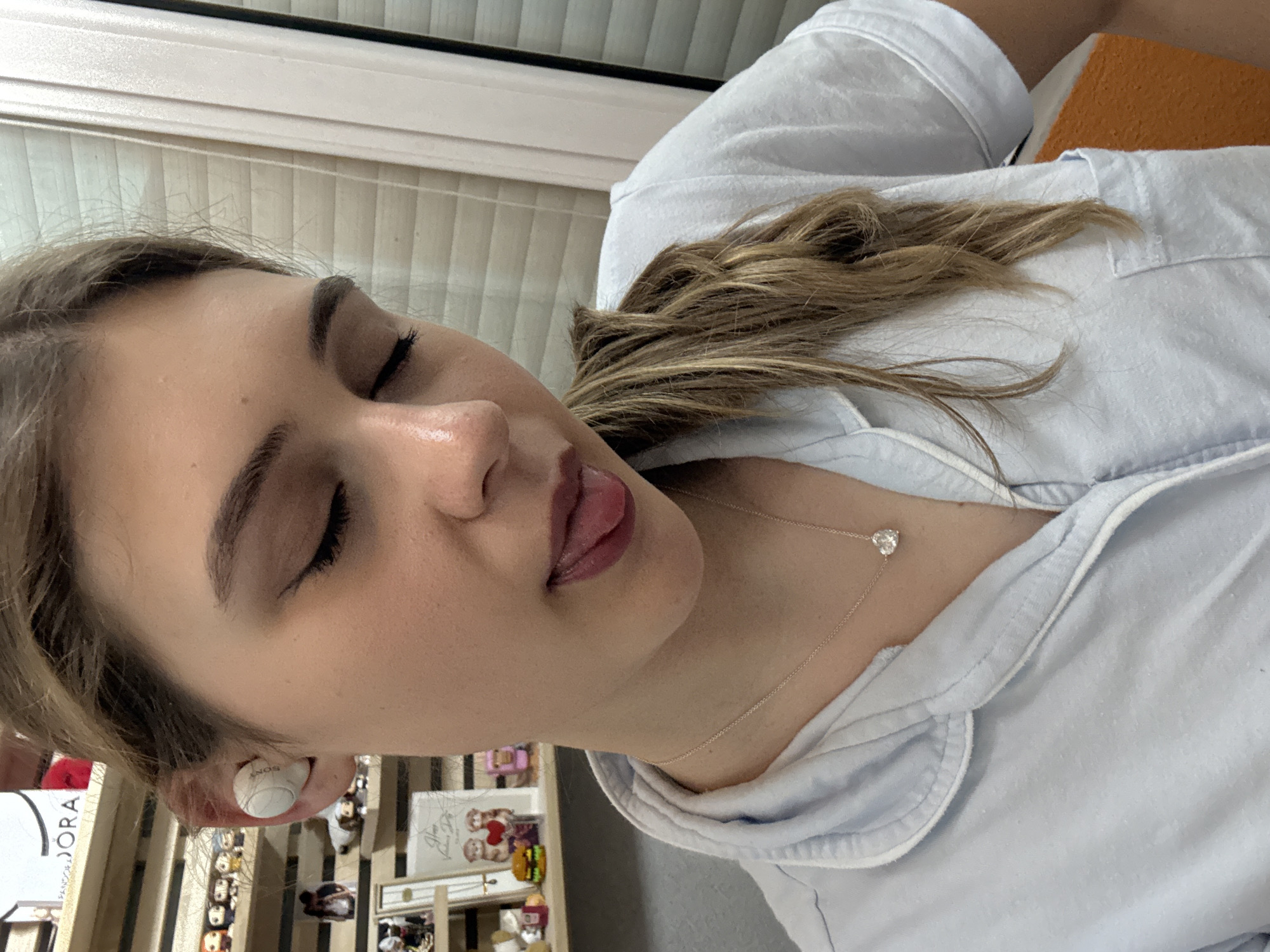 A close-up, canted shot depicts a young woman with light skin, her eyes closed, and her tongue slightly protruding from her mouth. She is wearing a light blue collared shirt or pajamas. A delicate silver necklace with a heart-shaped pendant is visible around her neck. She is wearing a white wireless earbud in her ear. In the blurred background, a display case with various small objects and miniature figures can be seen. Based on visual cues, the location cannot be confirmed to be in Panagyurishte, Bulgaria.