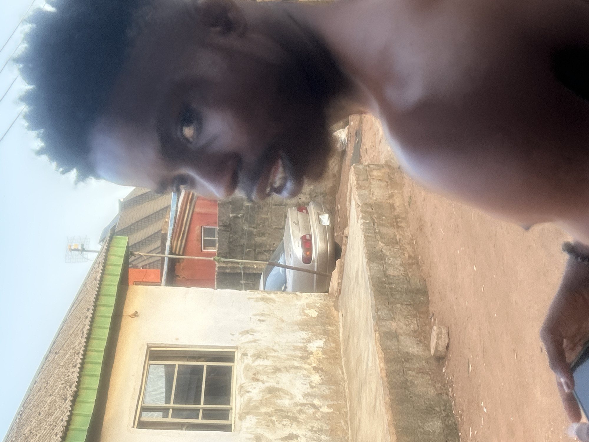 The image shows a person with dark skin and curly hair, likely male, taking a selfie at an angle. Behind him, there is a building with peeling paint and a window. The roof of the building has a green trim. In the background, a silver car is parked behind a wall. The ground is dirt. The city is Unknown, Unknown.