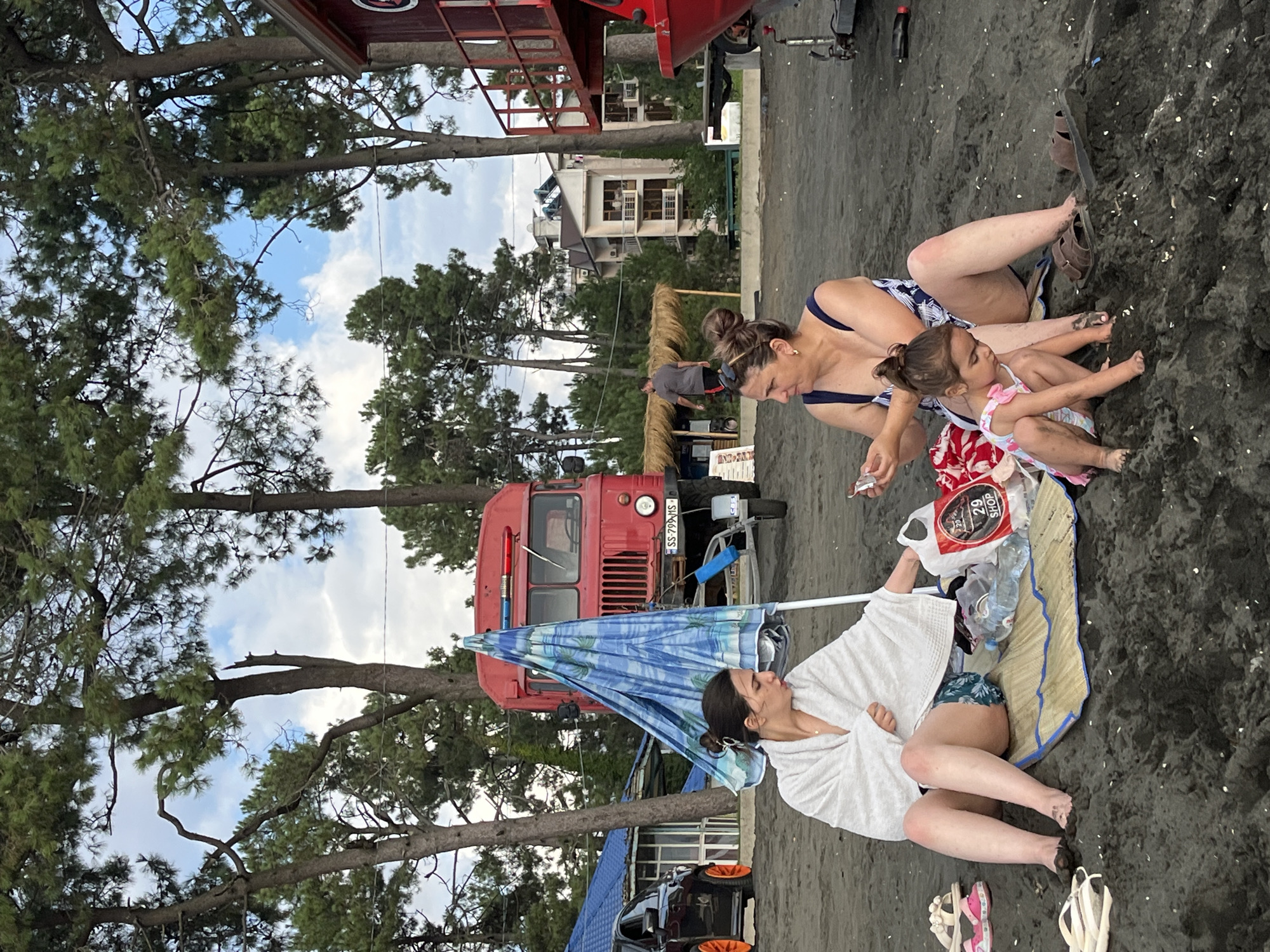 The image depicts a beach scene in Ureki, Georgia, with several people and objects on dark sand. In the foreground, two women and a young girl are seated on a mat. One woman, wrapped in a white towel, is looking down at items in front of her. The other woman, wearing a blue and white swimsuit, is holding a small object and looking towards the girl. The young girl, also in a swimsuit, is seated and appears to be playing in the sand.
Behind them, a blue and white beach umbrella is partially covering a red truck, possibly a vintage vehicle. A car with bright orange rims is parked to the left of the umbrella. Further in the background, there are trees, a building with multiple balconies, and the sea can be inferred to be nearby. The sky is partly cloudy. The overall atmosphere suggests a casual day at the beach.