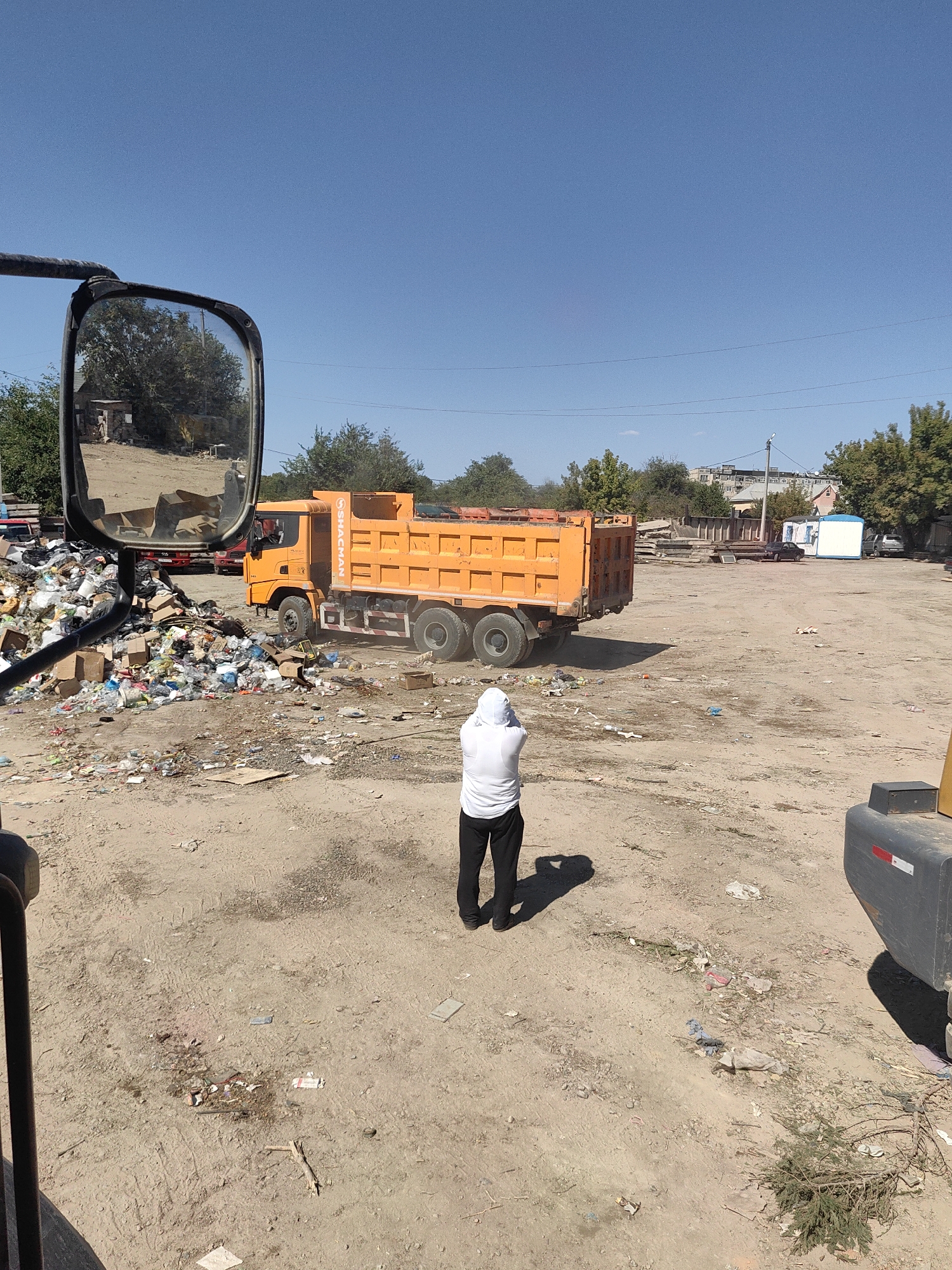 An outdoor, wide-angle, eye-level shot captures a scene of a landfill in Shymkent, Kazakhstan. The foreground is dominated by the reflective surface of a truck's side mirror, which shows a small, dilapidated shack and some trees. To the left of the mirror, a large pile of garbage spills out, a chaotic mix of plastic bottles, cardboard boxes, and other debris.
In the mid-ground, a bright orange dump truck, identified by the "SHACMAN" logo on its side, is parked. Its bed is raised, suggesting it may have just unloaded or is about to. A person wearing a white hooded jacket and black pants stands in the dirt, facing away from the camera and towards the dump truck, their shadow cast long on the ground. The ground is a mixture of dirt and scattered trash.
The background reveals more of the urban environment of Shymkent, with buildings, trees, and power lines under a clear blue sky. The overall impression is one of industrial waste disposal and the surrounding infrastructure.